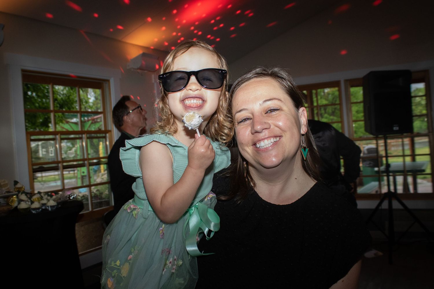 A child in a teal dress wears sunglasses while dancing at an indoor celebration.