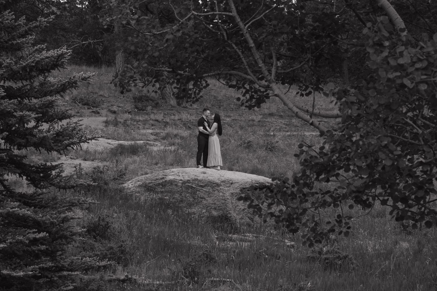 A black and white photo of a couple standing on a rock formation in a wooded setting.