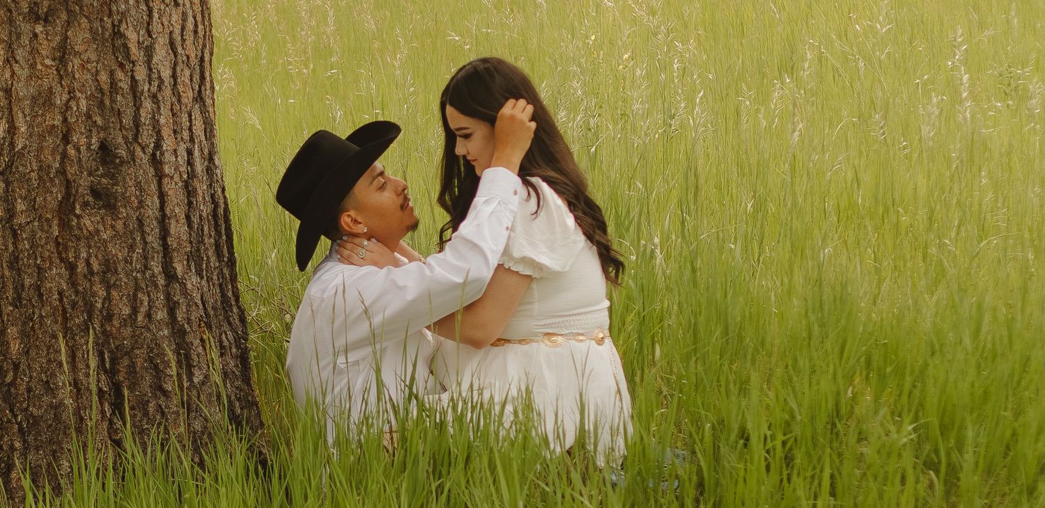 A romantic couple shares an intimate moment under a large tree in a sunlit meadow of tall grass.