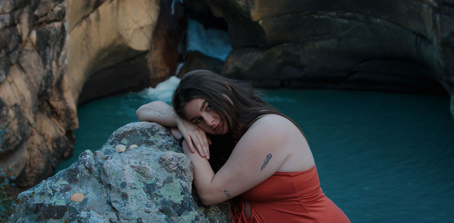 Person in red dress rests on rocky outcrop beside turquoise water in moody natural setting.