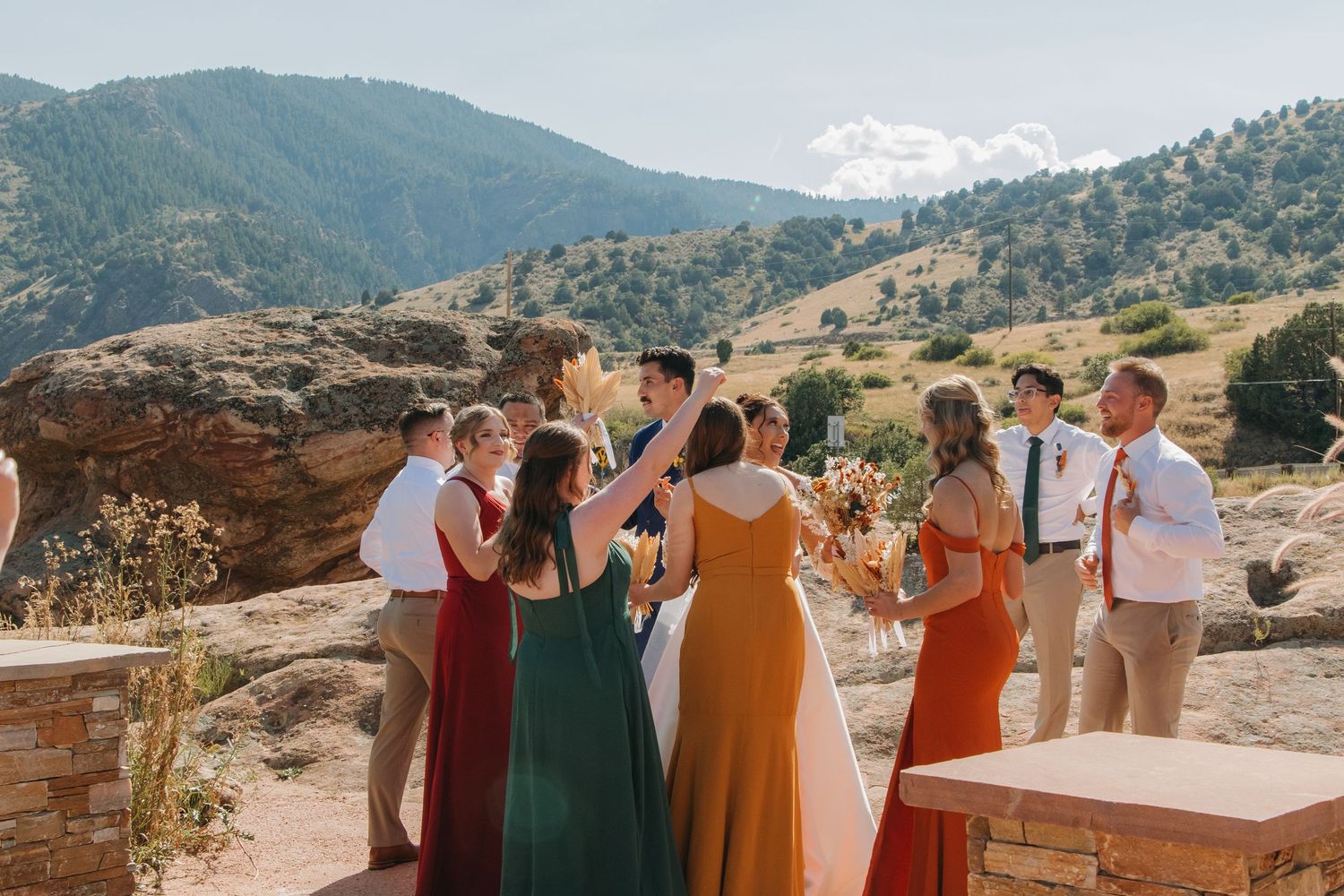 Wedding guests in colorful dresses stand together with a mountain landscape in the background.