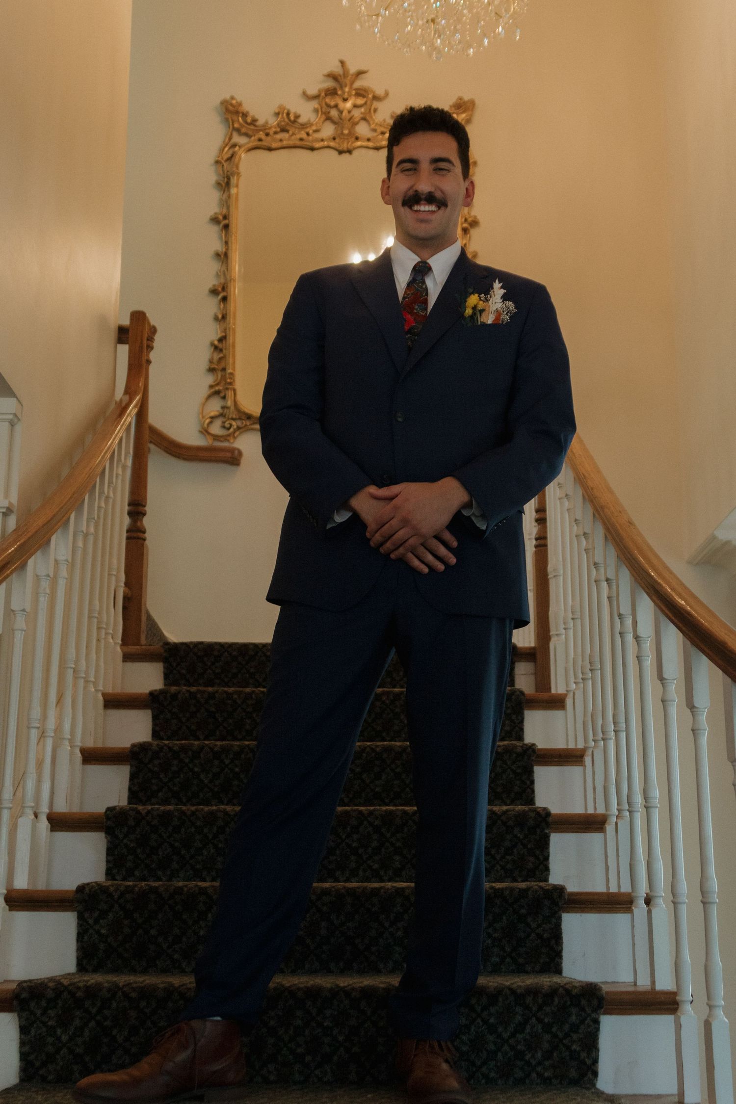 A person in a navy suit sits on carpeted stairs with ornate gold decor and white railings.