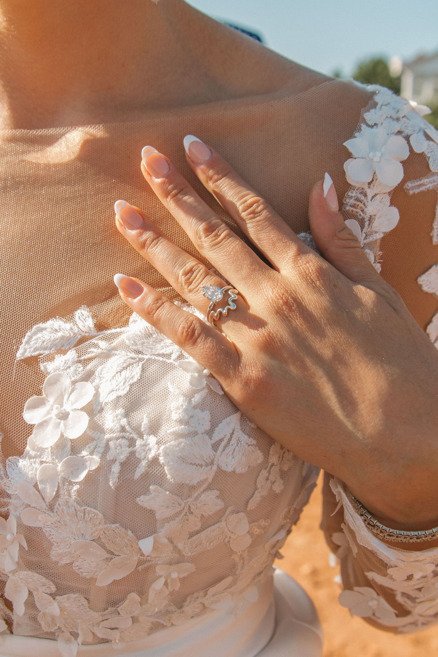 Close-up of delicate white lace wedding dress details and engagement ring in warm sunlight.