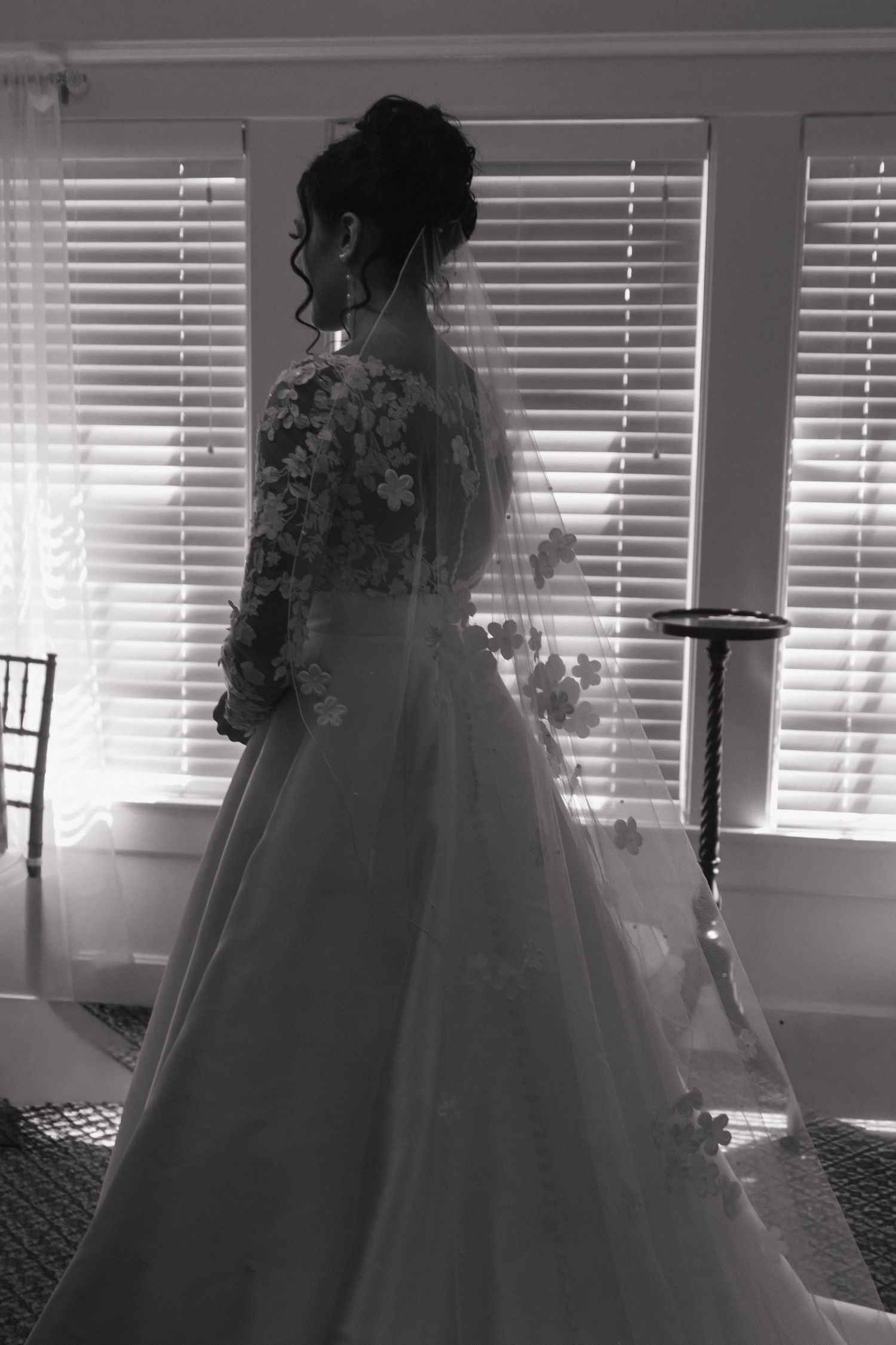 Silhouette of a bride in a flowing ball gown against window light with venetian blinds.