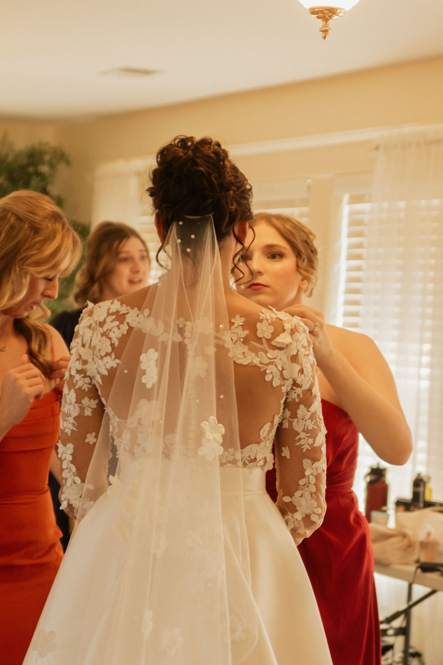 Bridesmaids in red dresses helping with a bride's intricate floral lace veil.