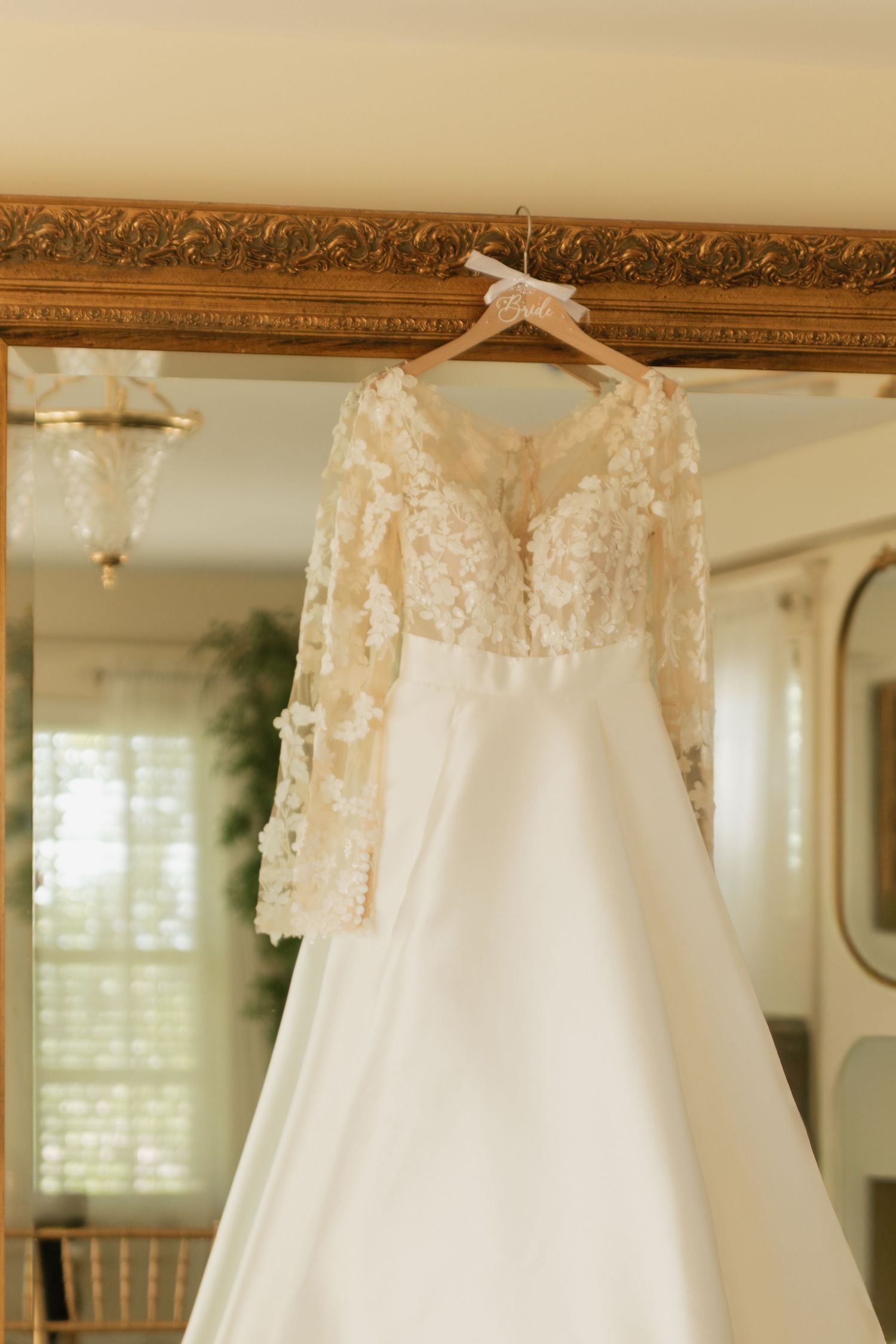 White wedding gown with delicate lace details hanging against a gold-framed mirror.