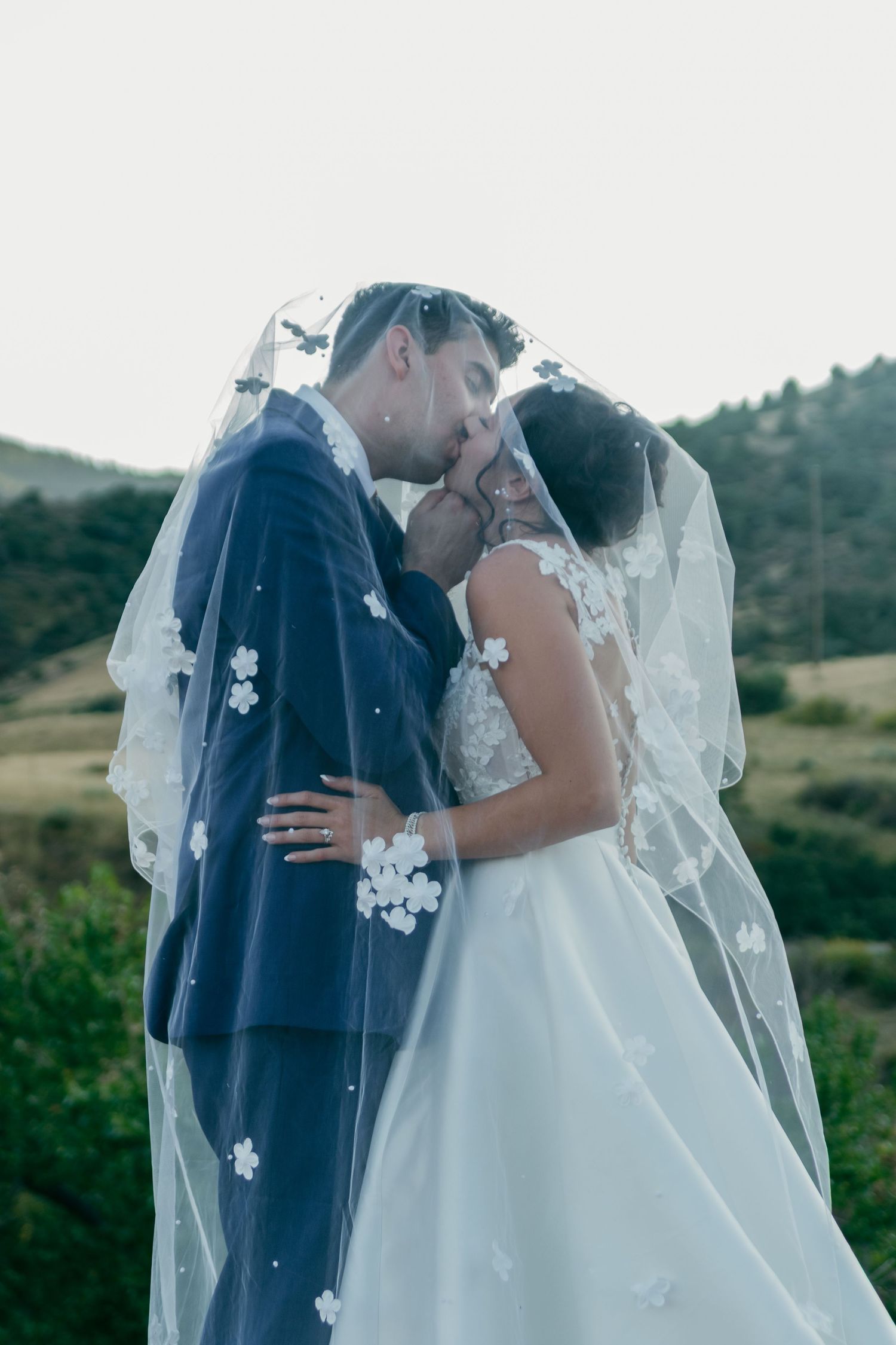 A couple shares an intimate kiss under a delicate floral veil against a mountain backdrop.