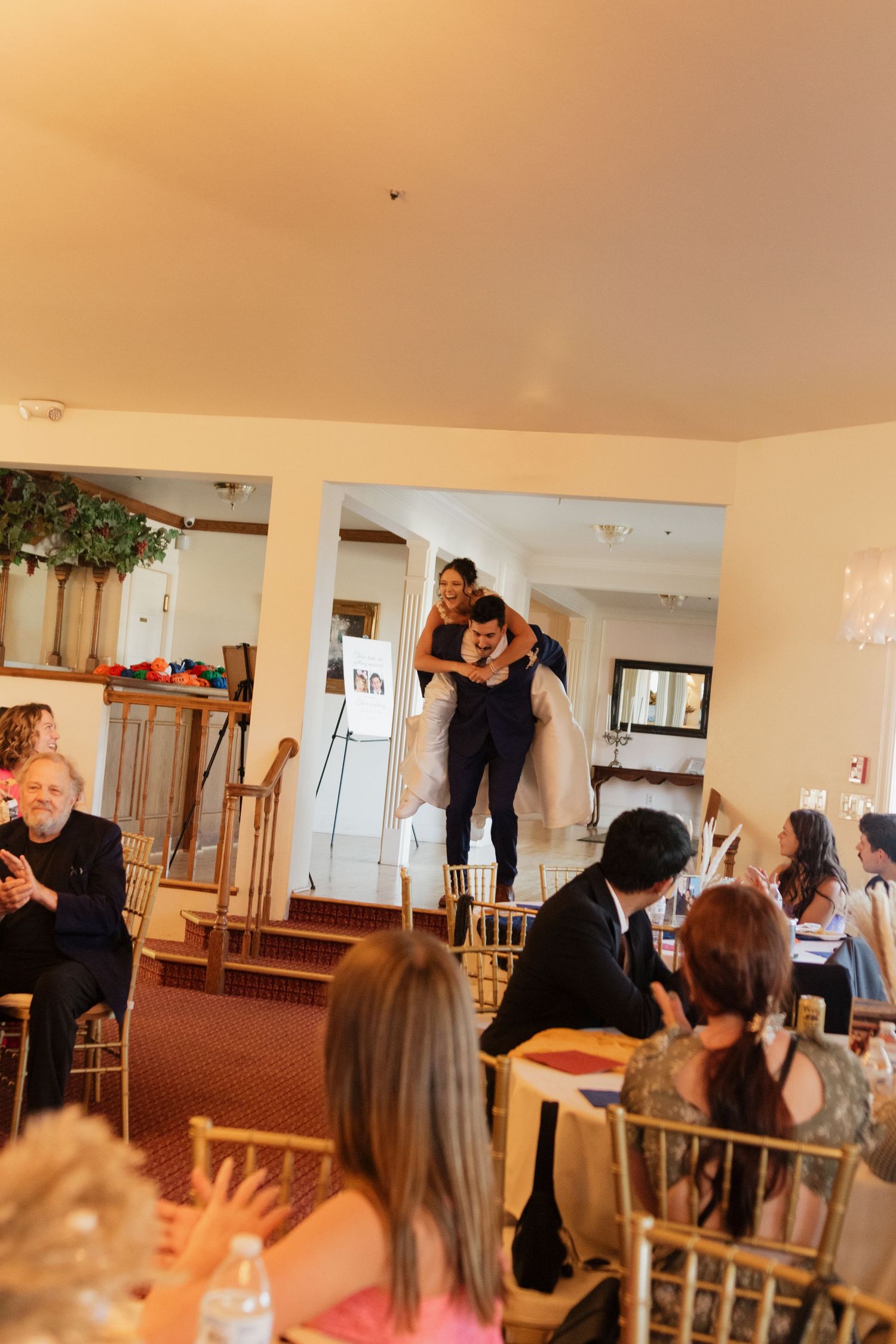 Wedding reception guests gathered in a bright venue with wooden chairs and warm lighting.