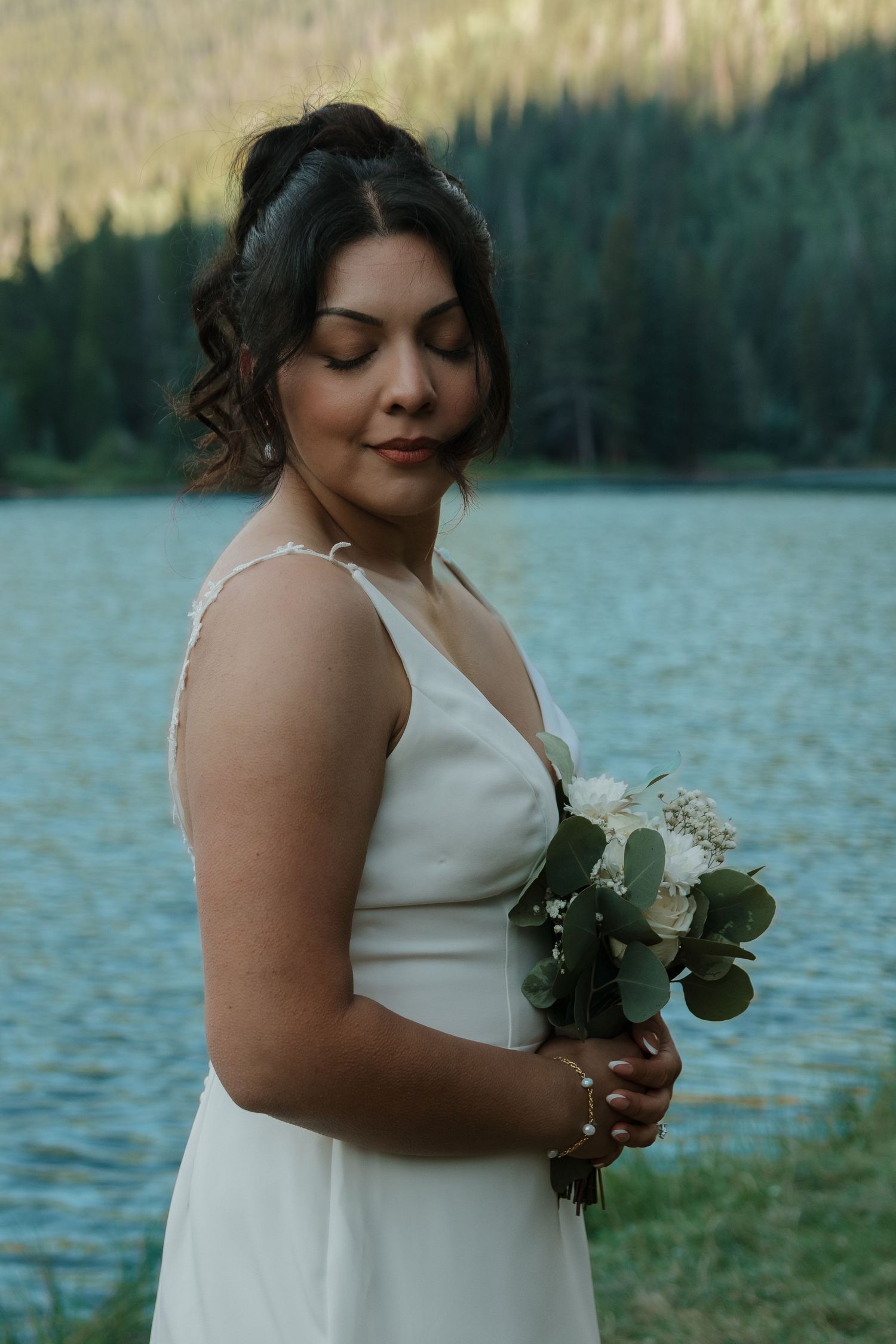 Person in white wedding dress holds bouquet beside mountain lake.