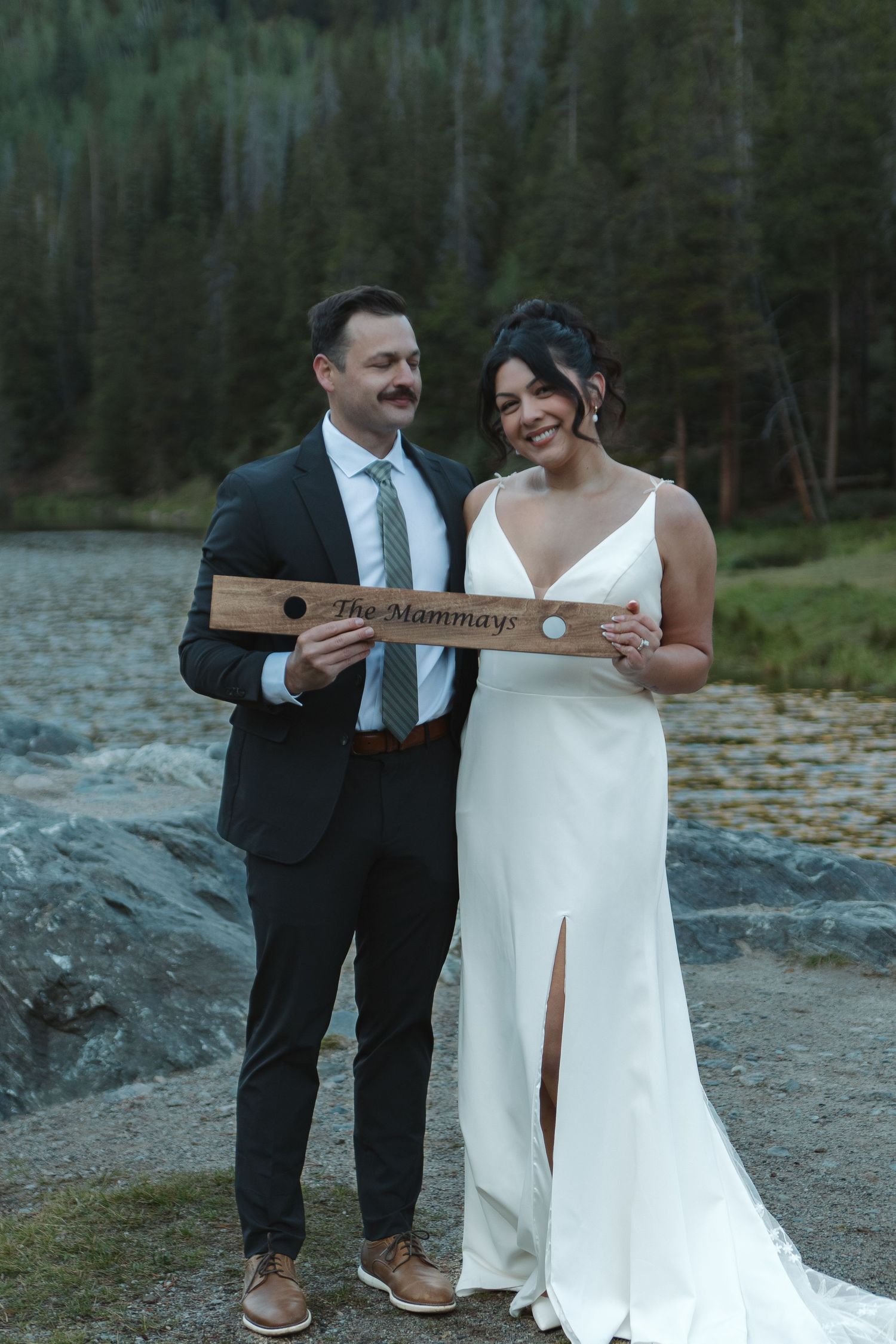 Wedding couple holds wooden sign reading 'the Hammonds' against forest and lake backdrop.