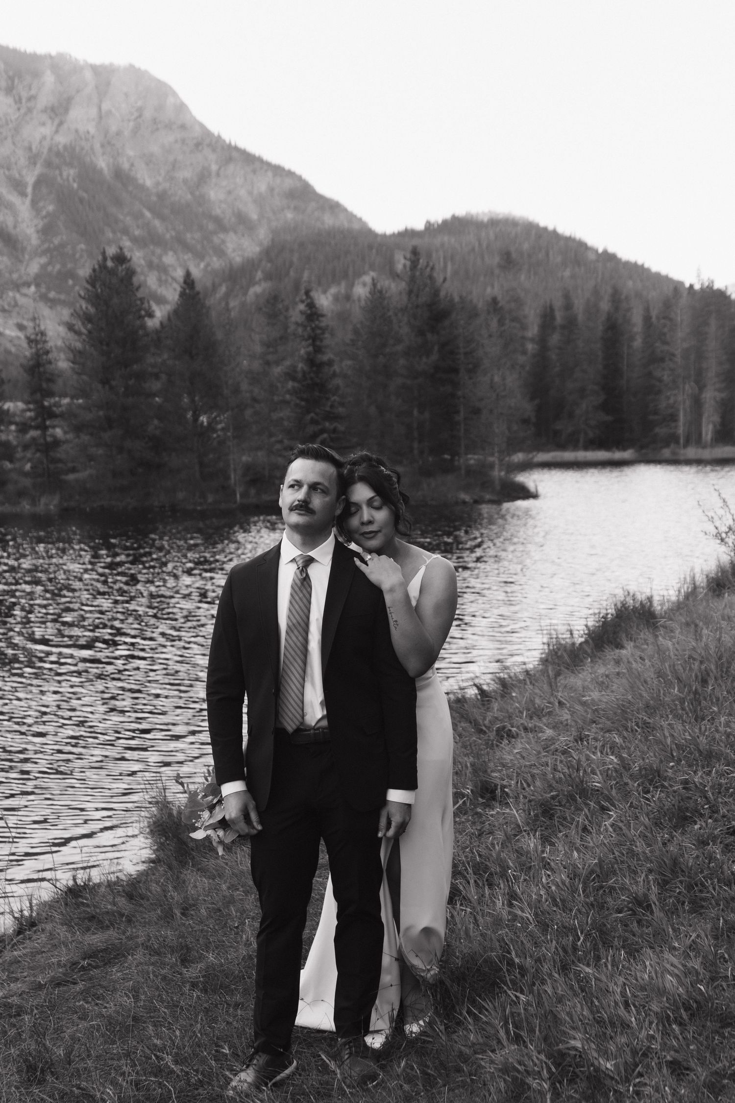 A couple poses together on a grassy shore with a mountain lake and evergreen forest backdrop.