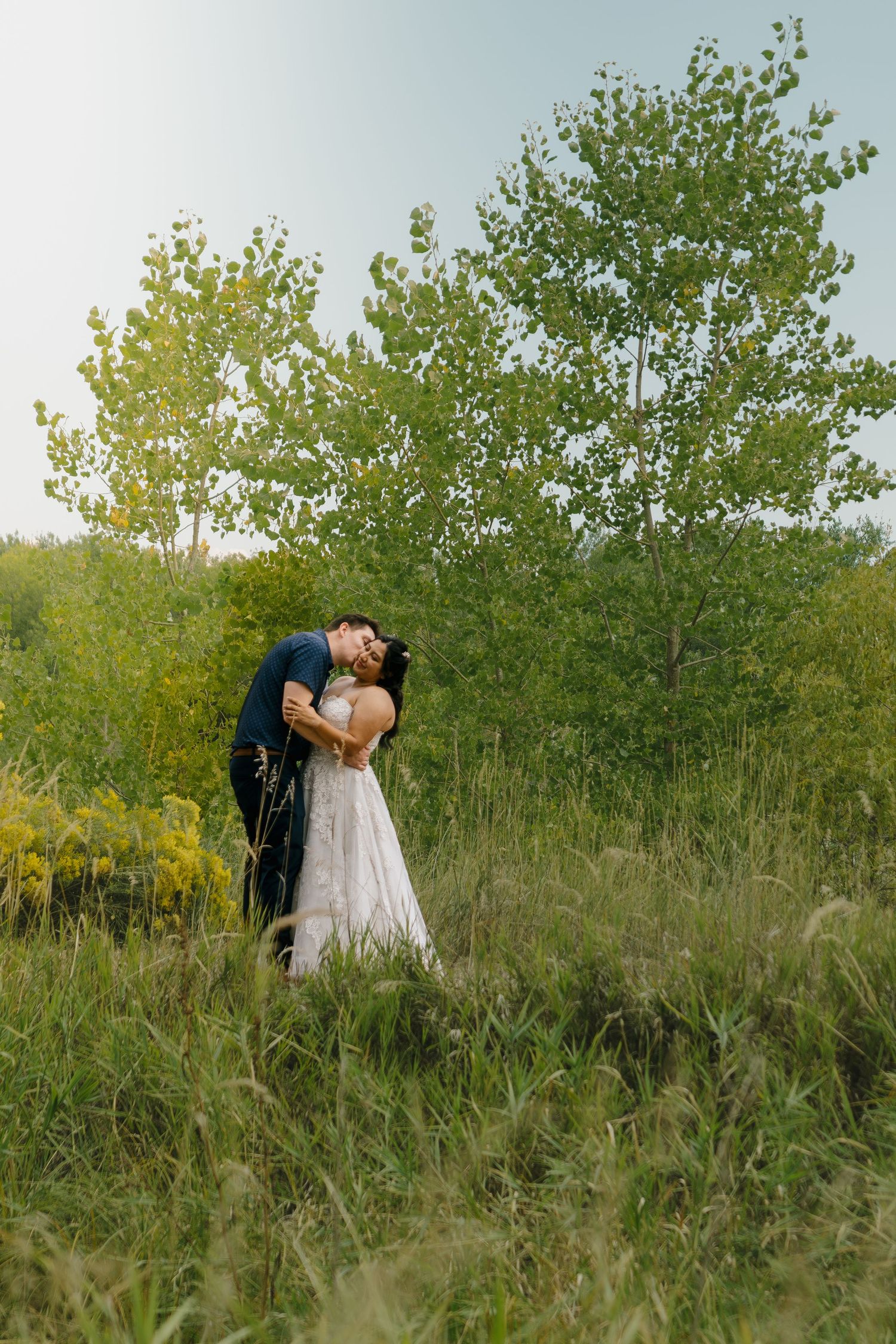 A couple shares intimate moments in a sunlit meadow surrounded by tall grass and trees during a golden hour photo session.