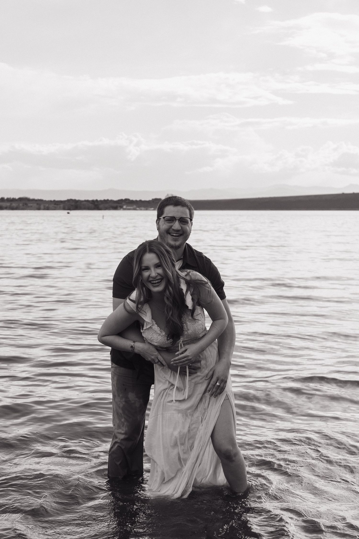 Black and white photo of a joyful couple laughing and embracing while standing in a lake fully clothed.