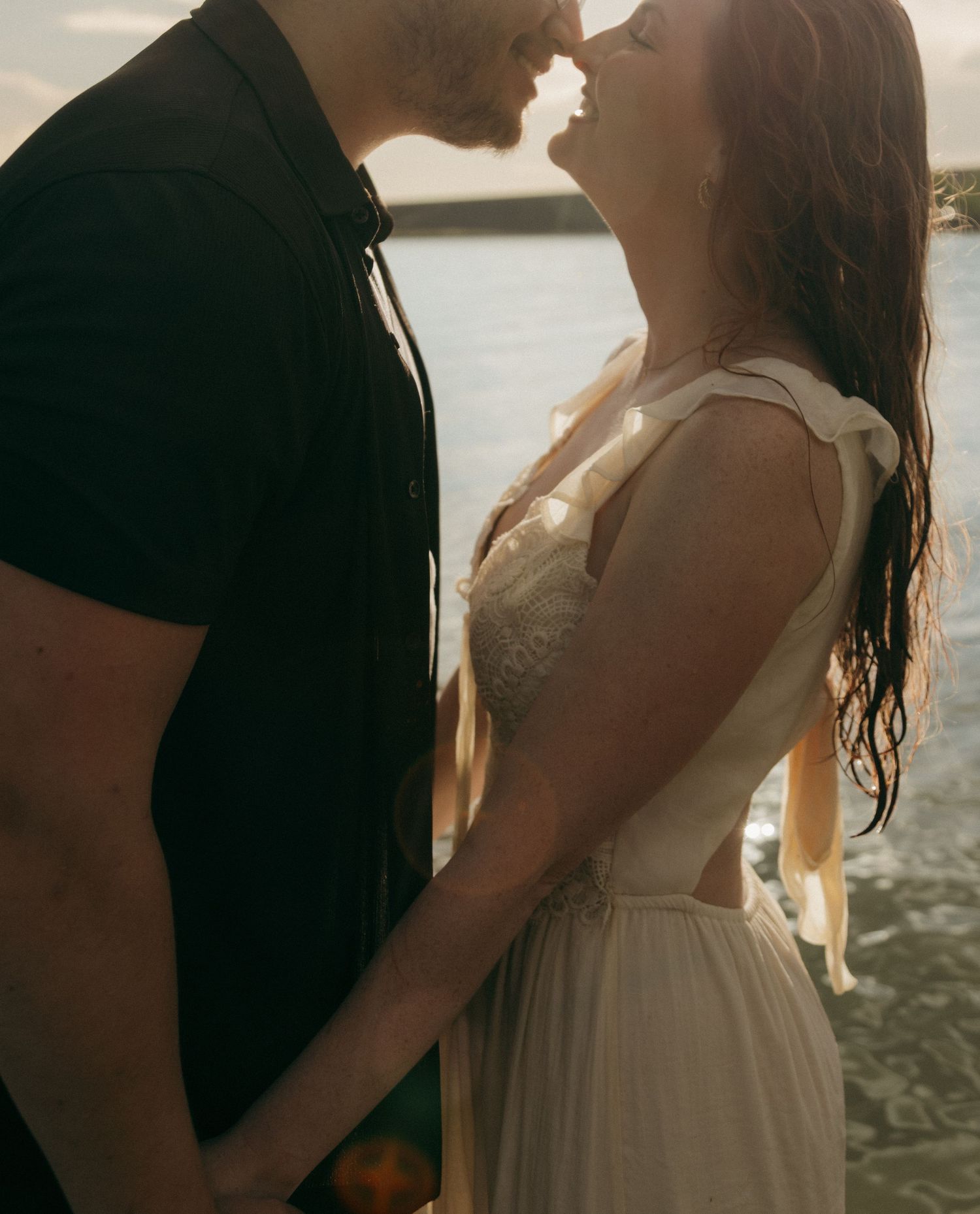 Close-up of couple about to kiss while holding hands near sunlit lake, woman in white dress.
