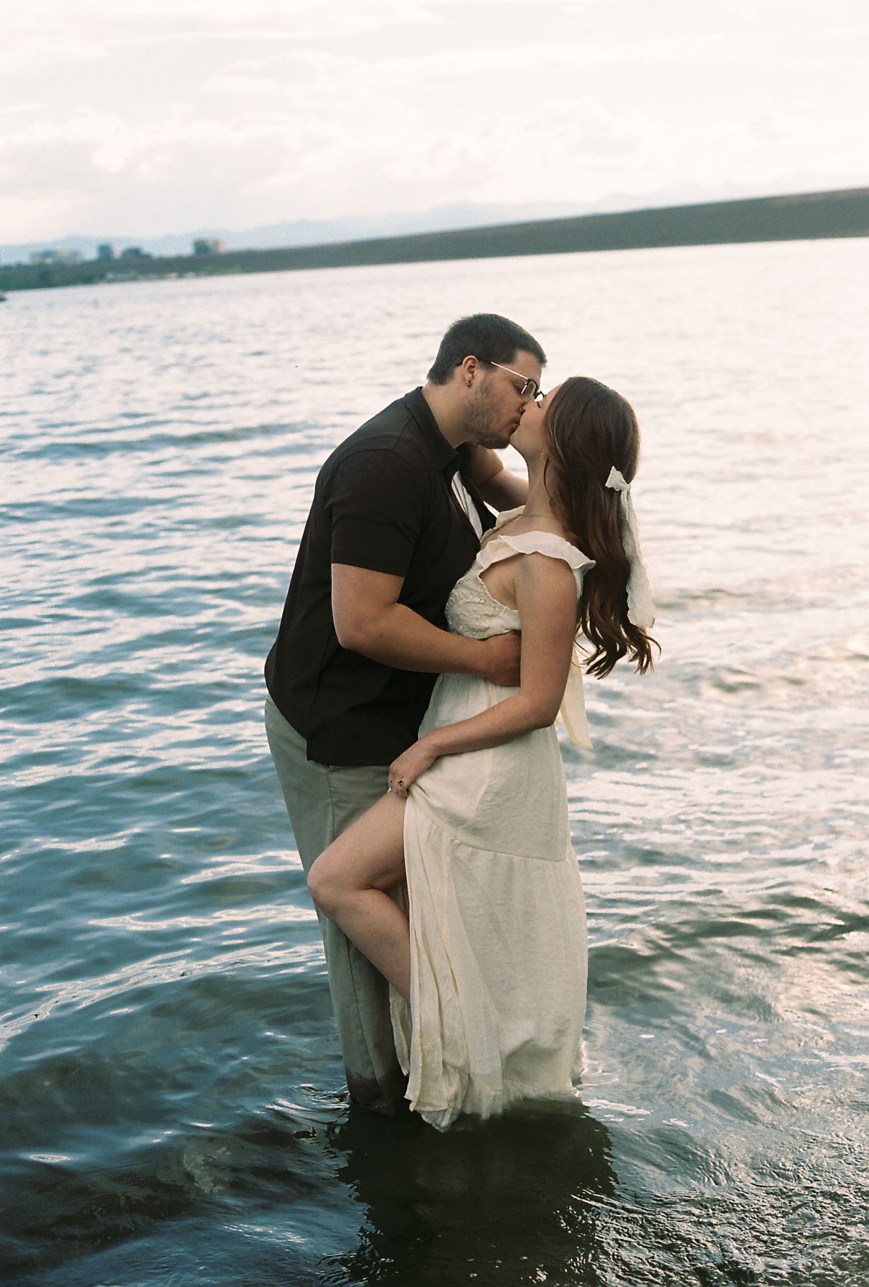 Couple sharing a kiss while standing waist-deep in a calm lake at dusk.