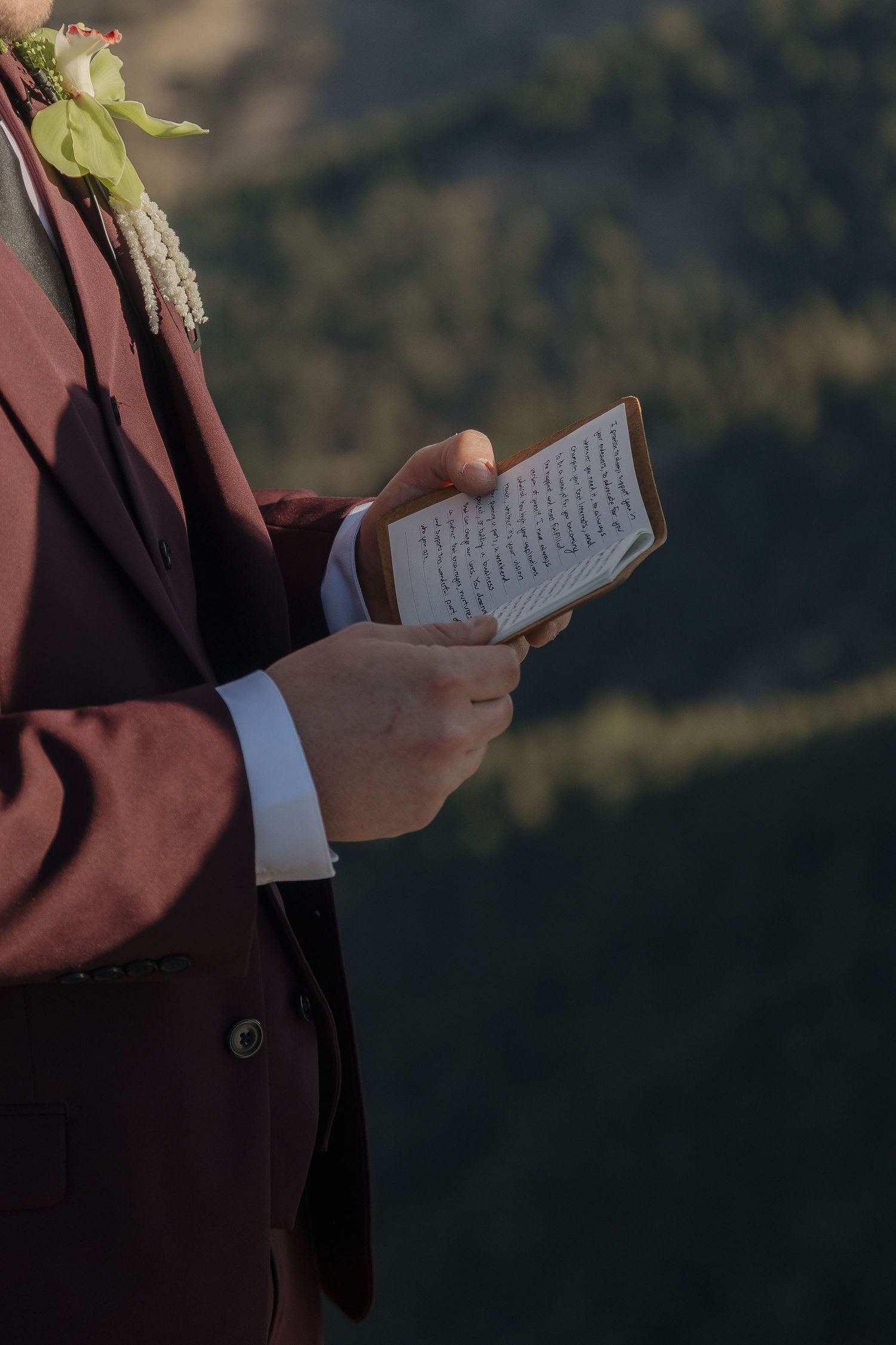 Groom in burgundy suit reads handwritten vows from a small notebook with mountain backdrop.