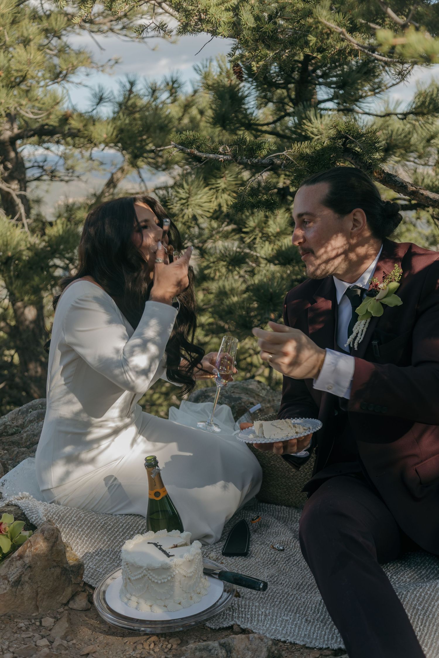 Bride and groom laughing while sharing cake and champagne at outdoor mountain elopement picnic.