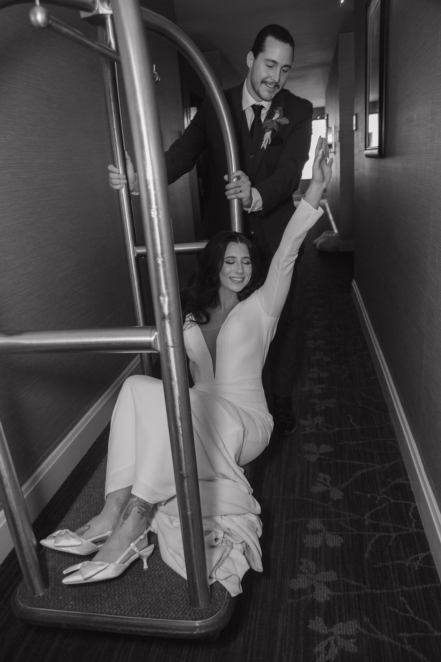 Black and white photo of playful bride and groom posing with a luggage cart in a hotel hallway.