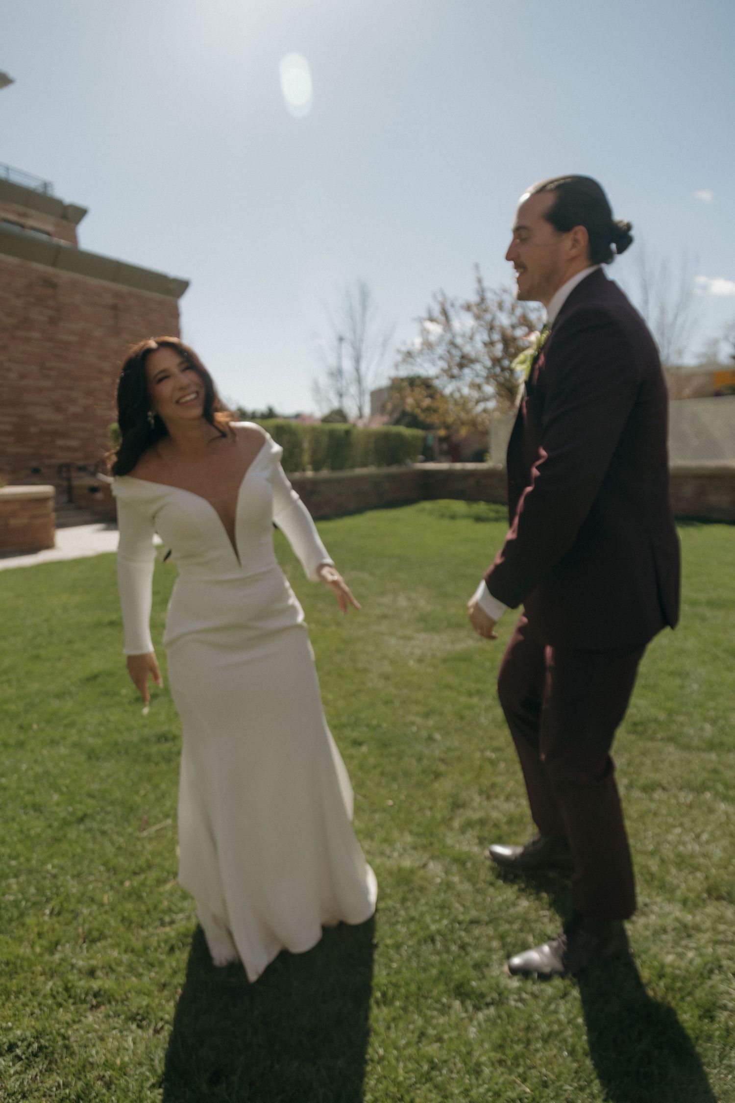 Bride in off-shoulder white gown and groom in burgundy suit laughing together on a sunny lawn.