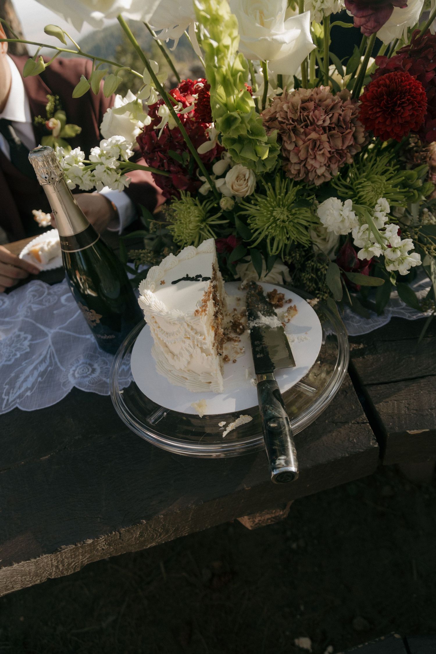 Close-up of a sliced white wedding cake on a clear stand beside a champagne bottle and lush floral centerpiece.
