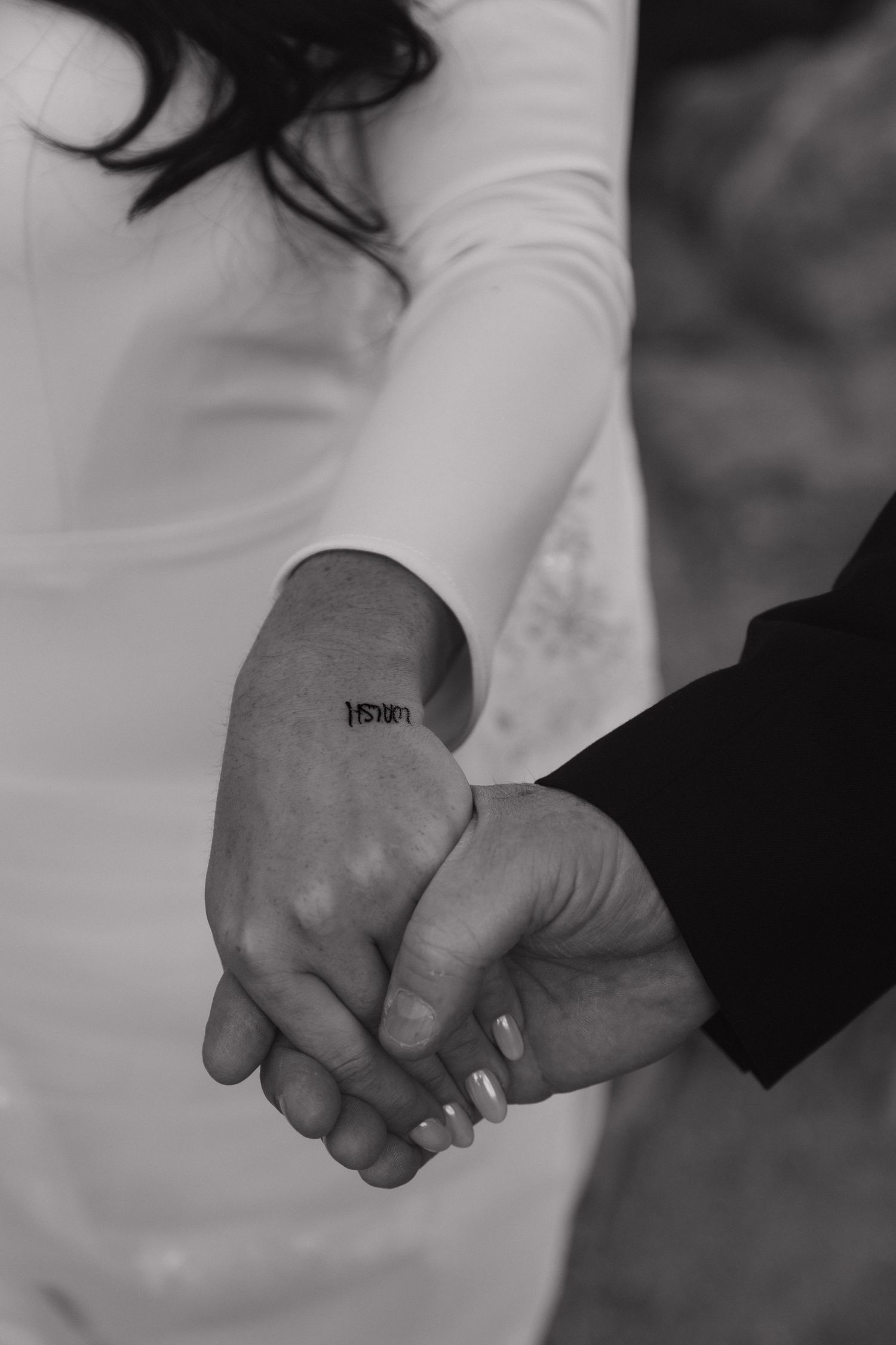 Black and white close-up of bride and groom holding hands showing wrist tattoo.
