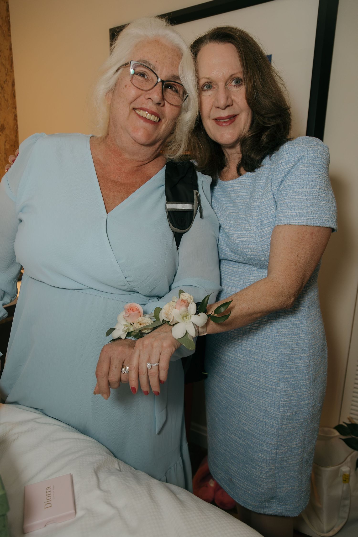 Two women in light blue dresses pose together wearing floral wrist corsages at a wedding celebration.