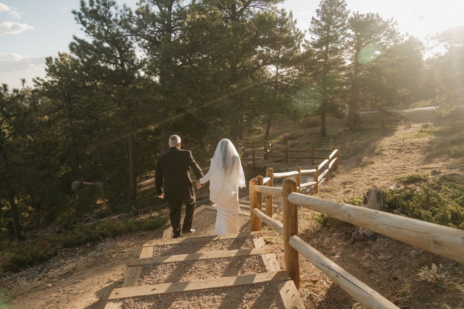 Bride in white gown and veil walking up wooden staircase with escort through pine forest.
