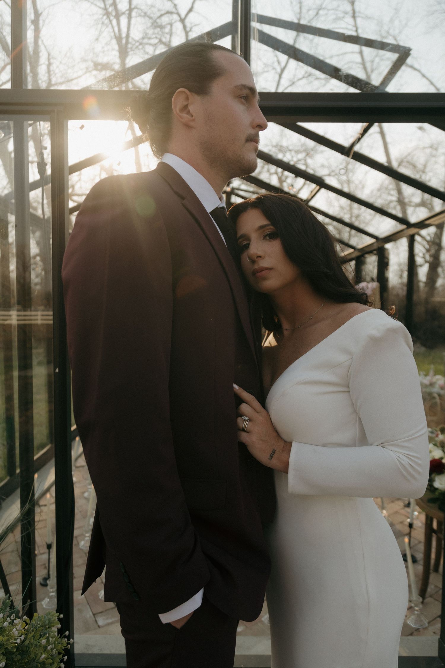 Bride in white gown leans on groom in burgundy suit with sunlight through greenhouse.