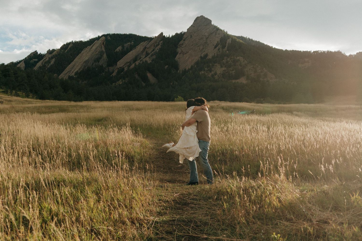 Man lifts woman in white dress on sunlit trail with iconic Boulder Flatirons mountains behind them.