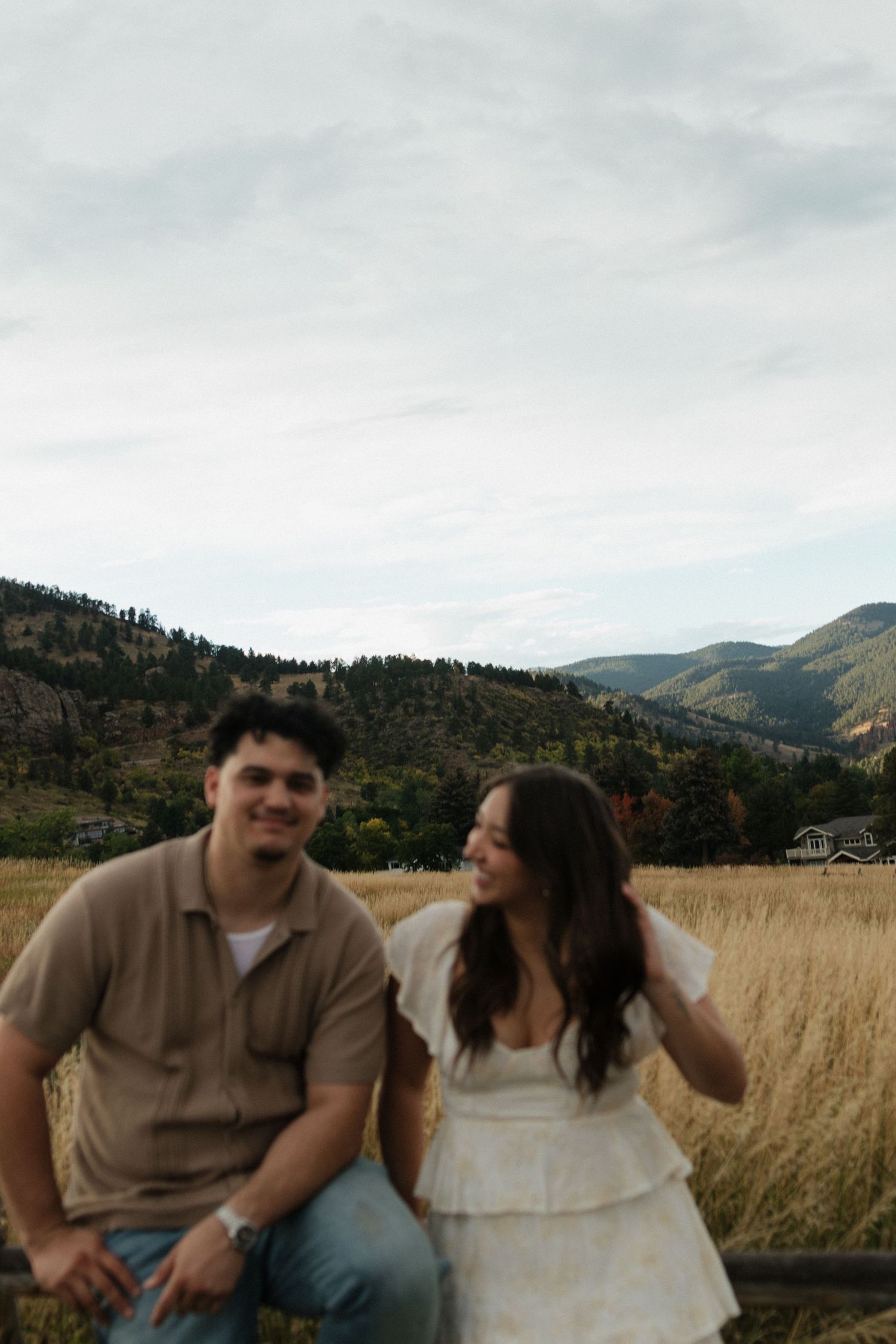 Smiling couple sitting in golden field with mountain landscape backdrop on cloudy day.