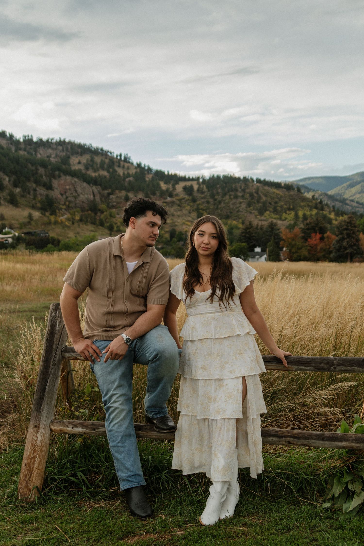 Couple posing by rustic wooden fence with mountain valley and golden field in background.