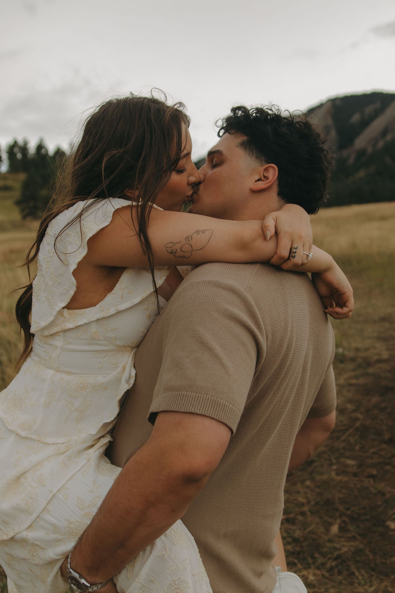 Couple sharing a romantic kiss in a golden field with mountain backdrop during outdoor engagement session.