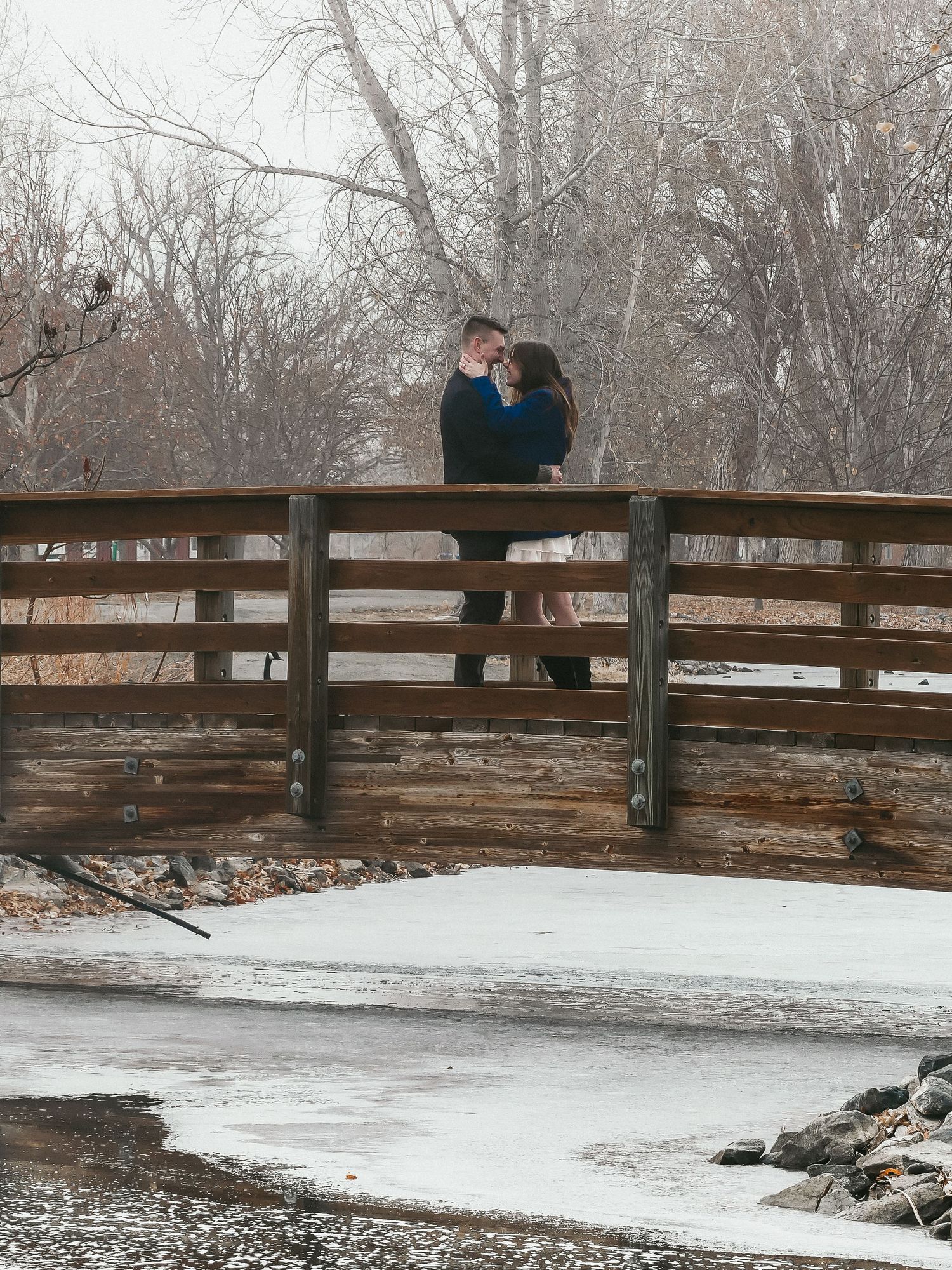 A couple standing close together on a bridge over a frozen stream.