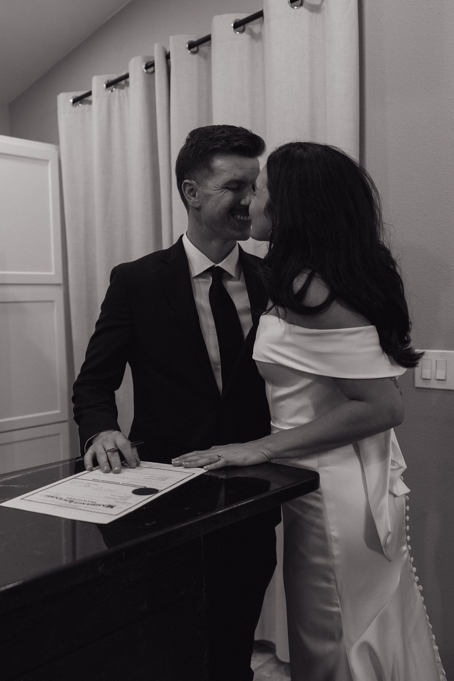 Black and white photo of bride and groom sharing a kiss while signing their marriage certificate.