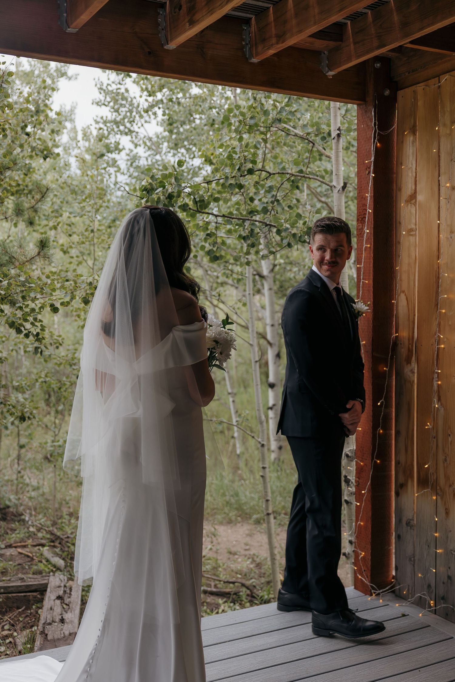 Groom smiling as bride in white gown and veil turns for first look on rustic wooden deck.