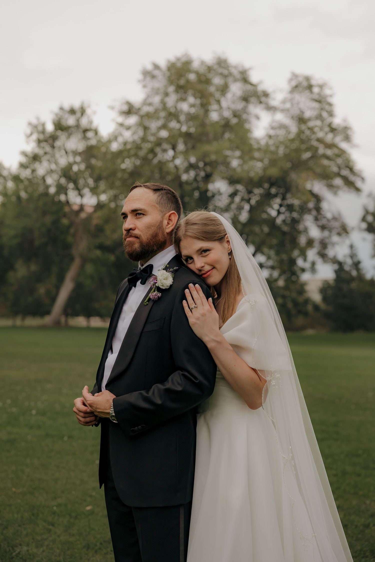 Bride in white gown and veil embraces groom in black tuxedo in lush green park setting.