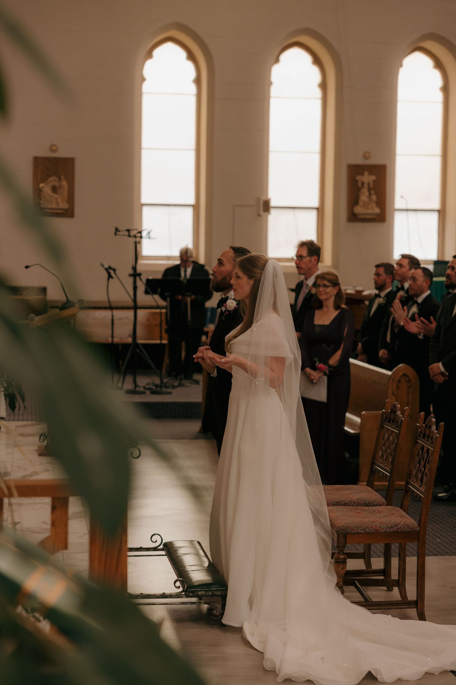 Bride in white gown and veil stands at church altar during wedding ceremony with guests and wedding party.