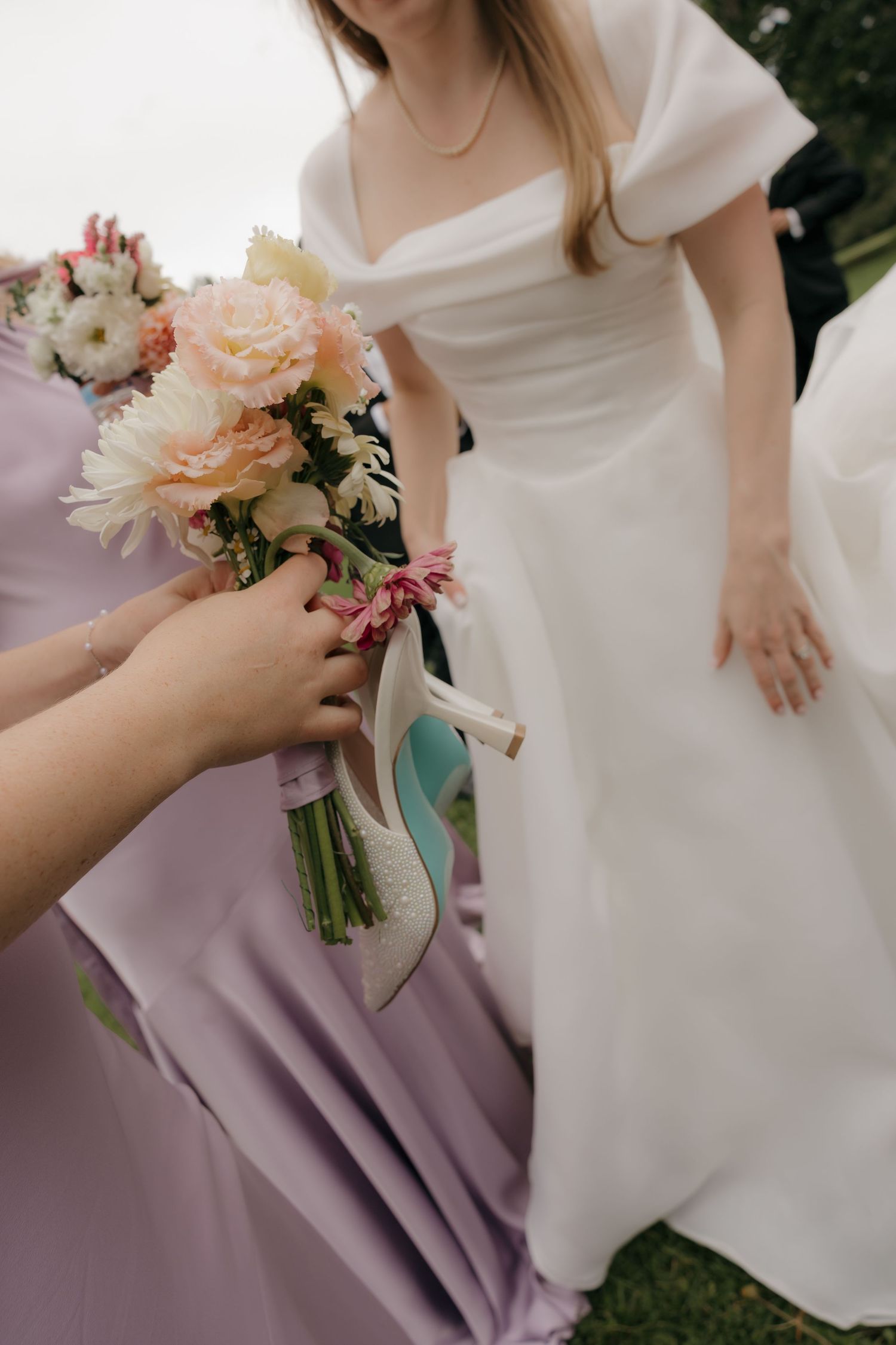 Bridesmaid in lavender dress handing bride a bouquet of pink and peach flowers outdoors.