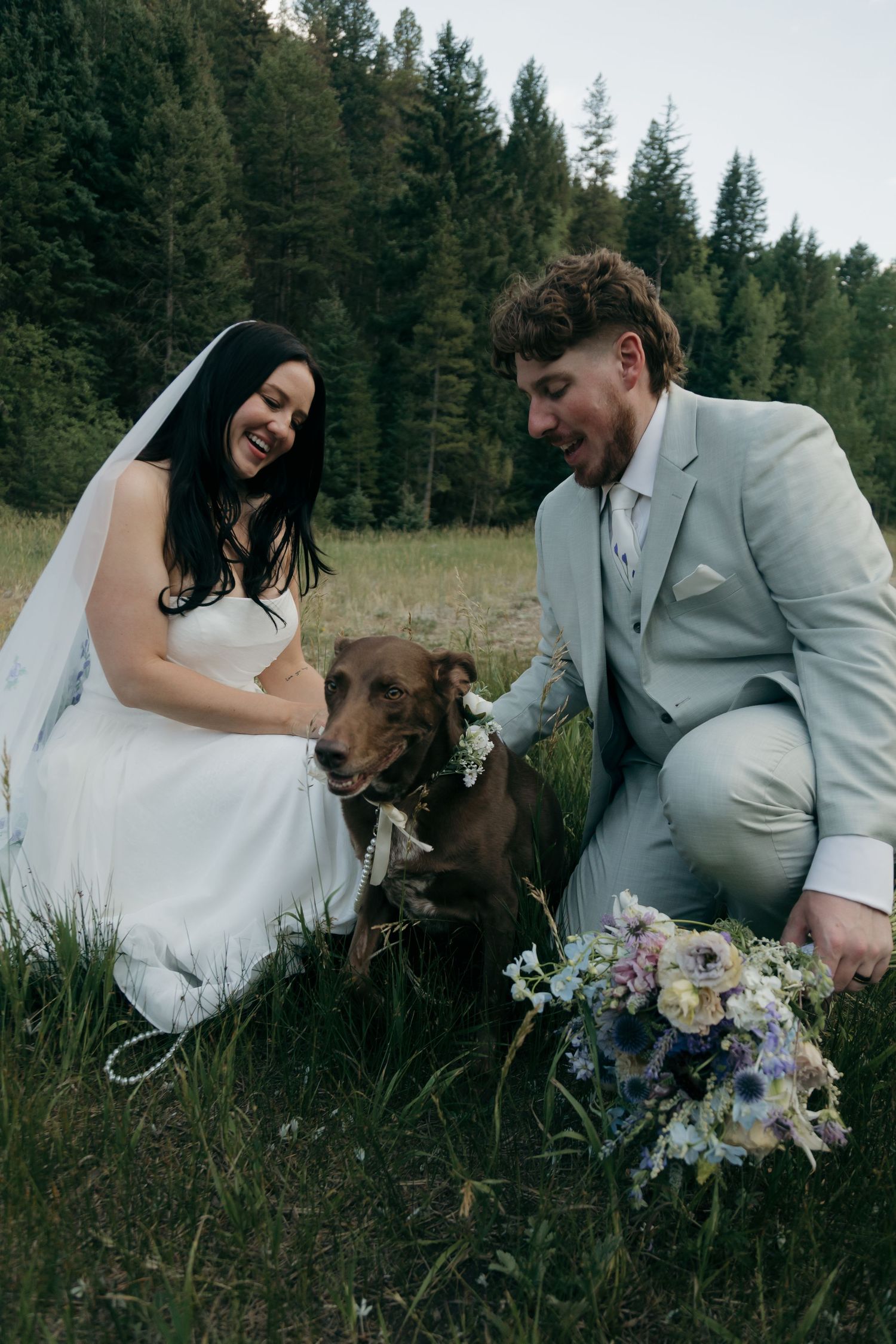 Laughing bride and groom in gray suit kneel in grass petting their flower-crowned chocolate Labrador.