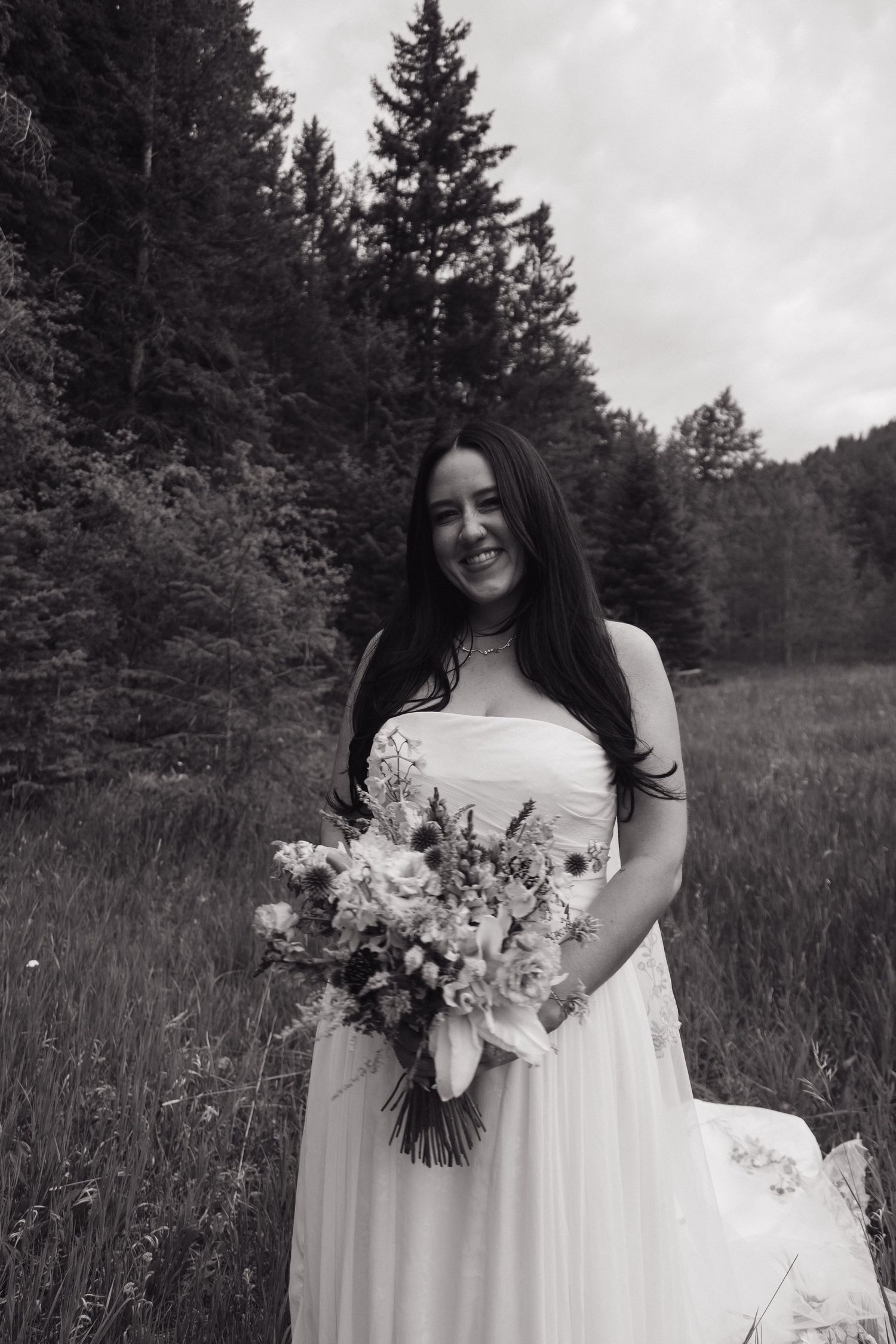 Black and white portrait of smiling bride holding a wildflower bouquet in a forest meadow.