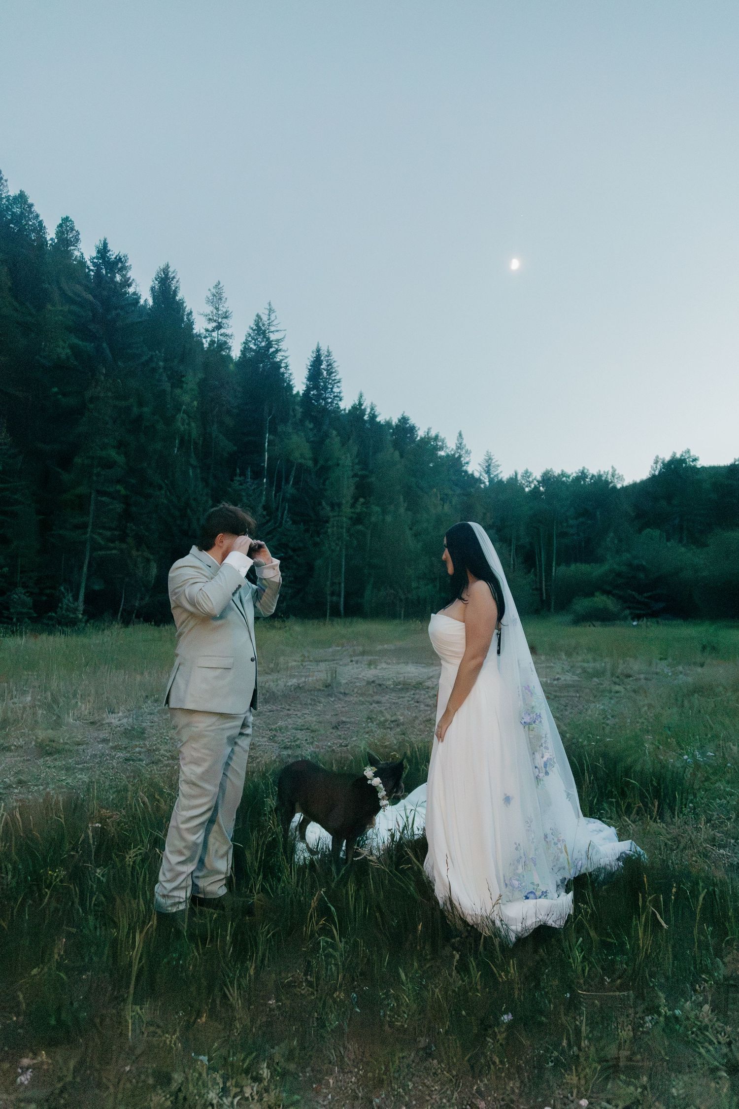 Groom photographs bride in white gown with dog in moonlit forest meadow at dusk.