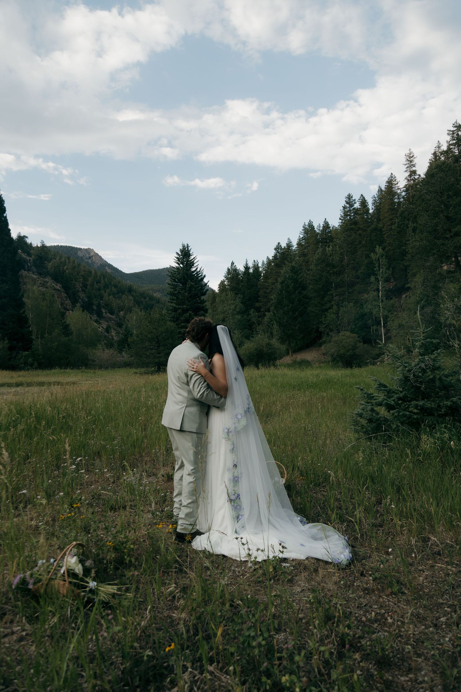 Wedding couple shares intimate embrace in a scenic mountain meadow with evergreen forest backdrop.