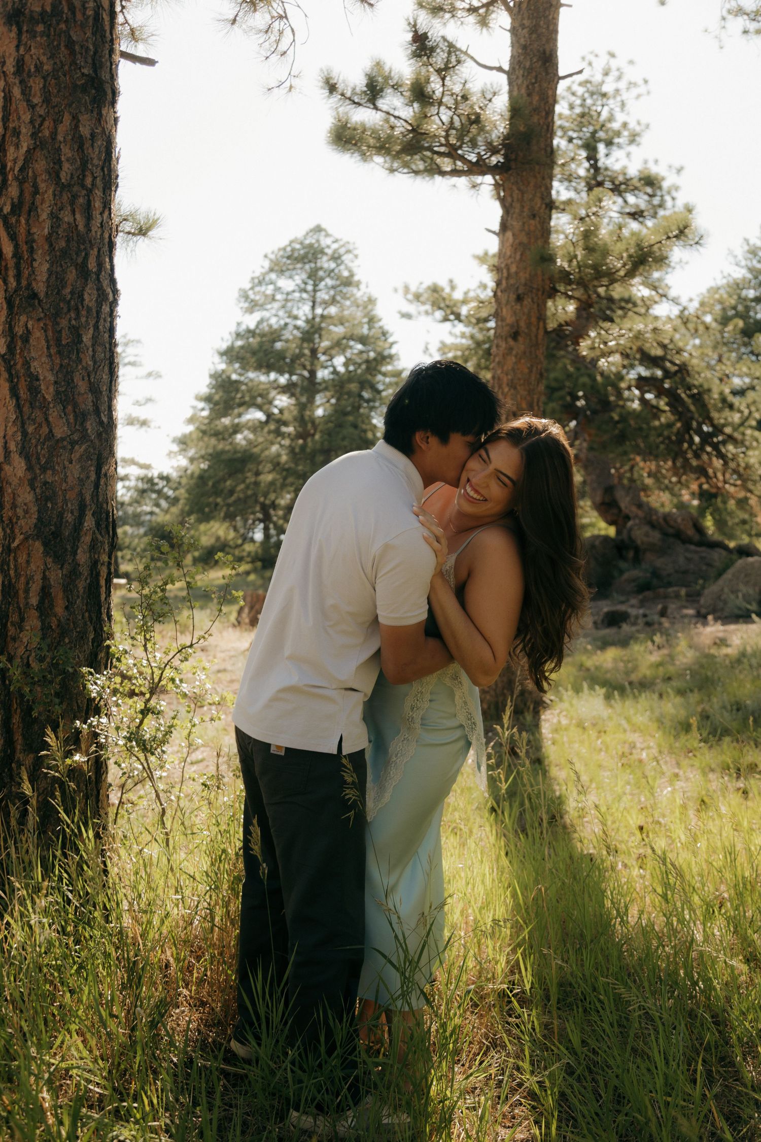 A couple embraces romantically in a sunlit forest clearing, the woman smiling as her partner kisses her cheek.