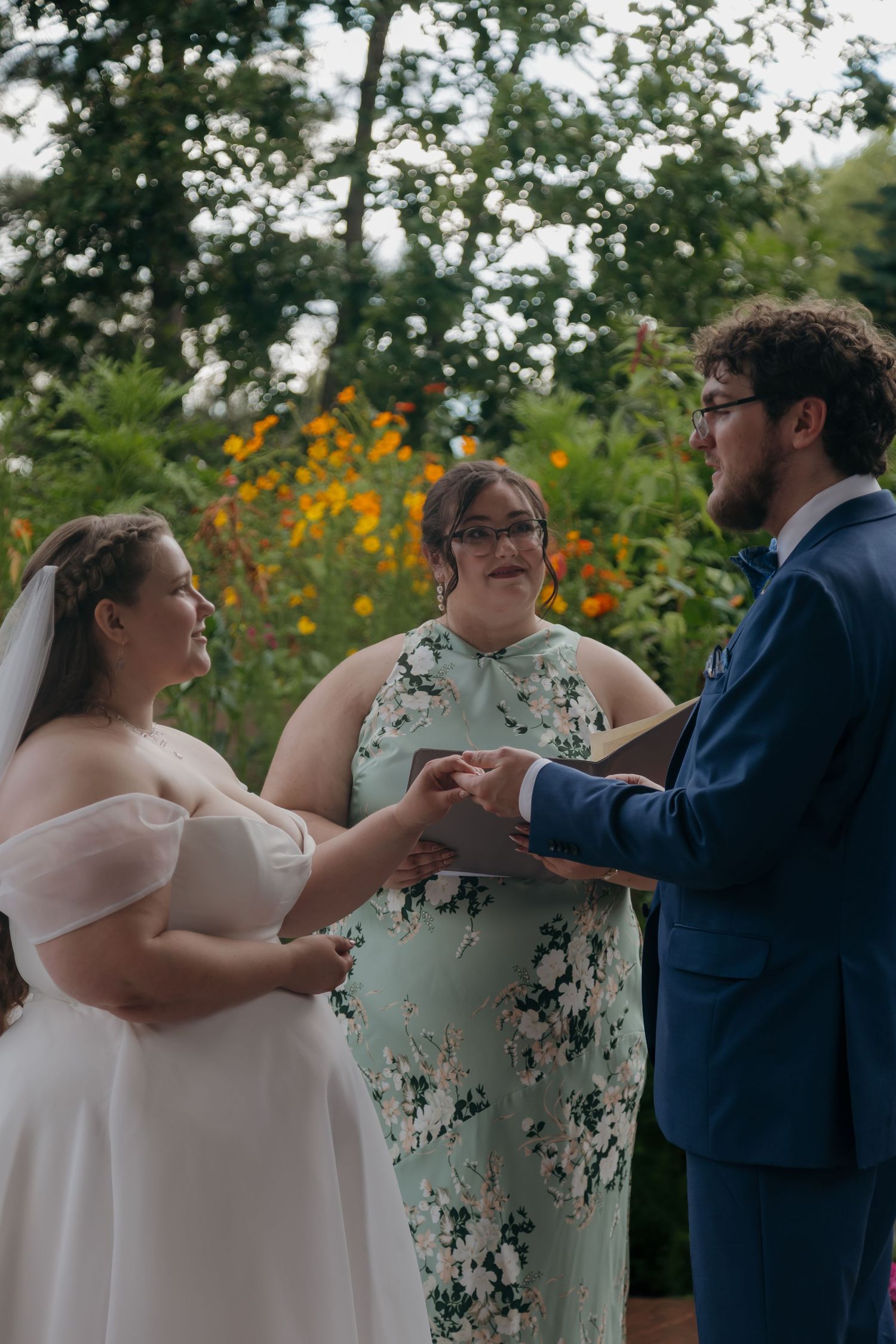 Bride and groom exchanging rings during outdoor garden wedding ceremony with officiant in floral dress.