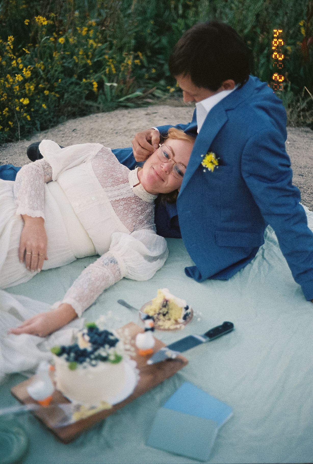 Bride in lace dress and groom in blue suit relaxing on a blanket at an outdoor wedding picnic.