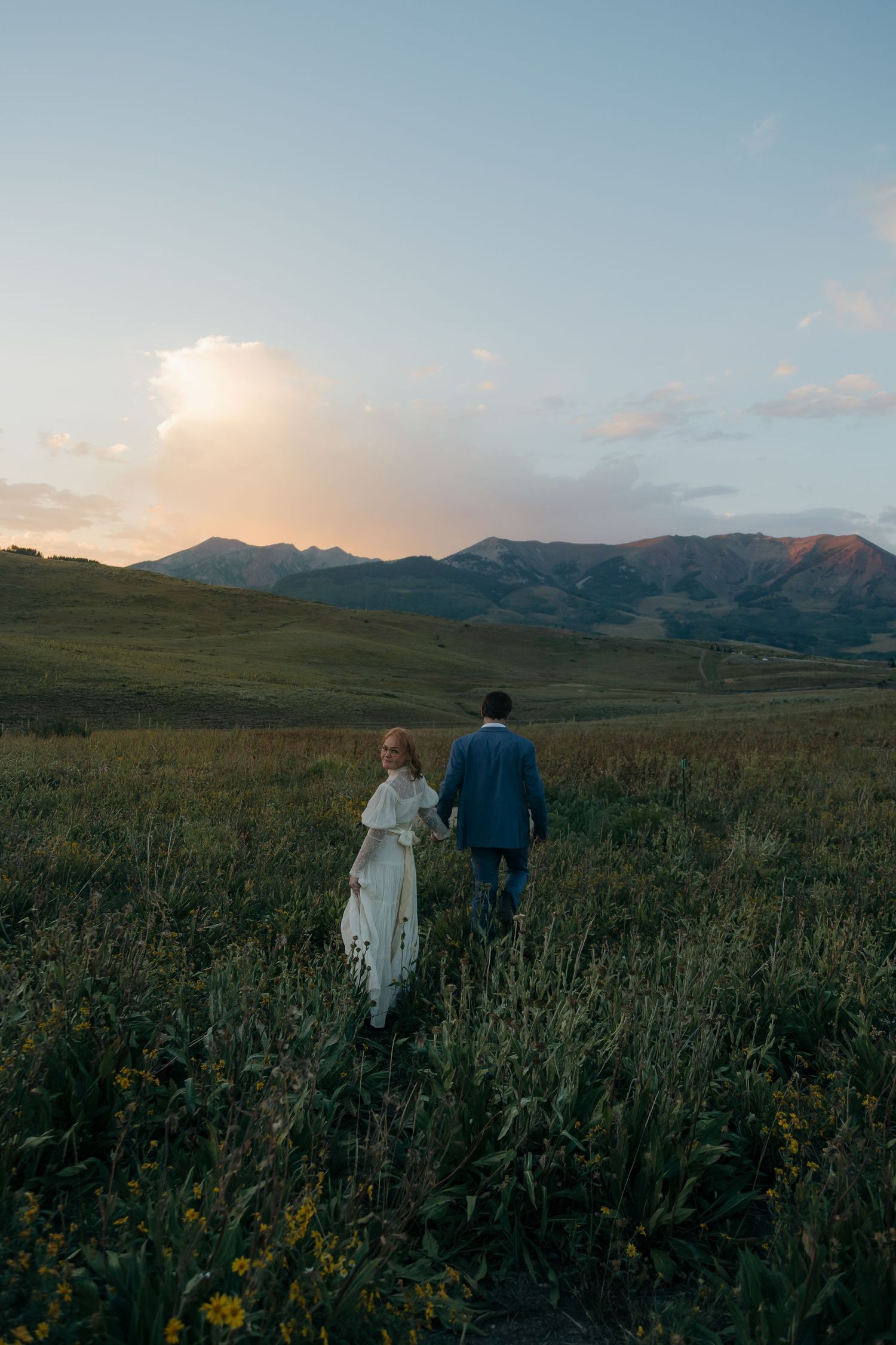 Bride and groom walk hand in hand through a wildflower meadow at sunset with mountain views behind them.