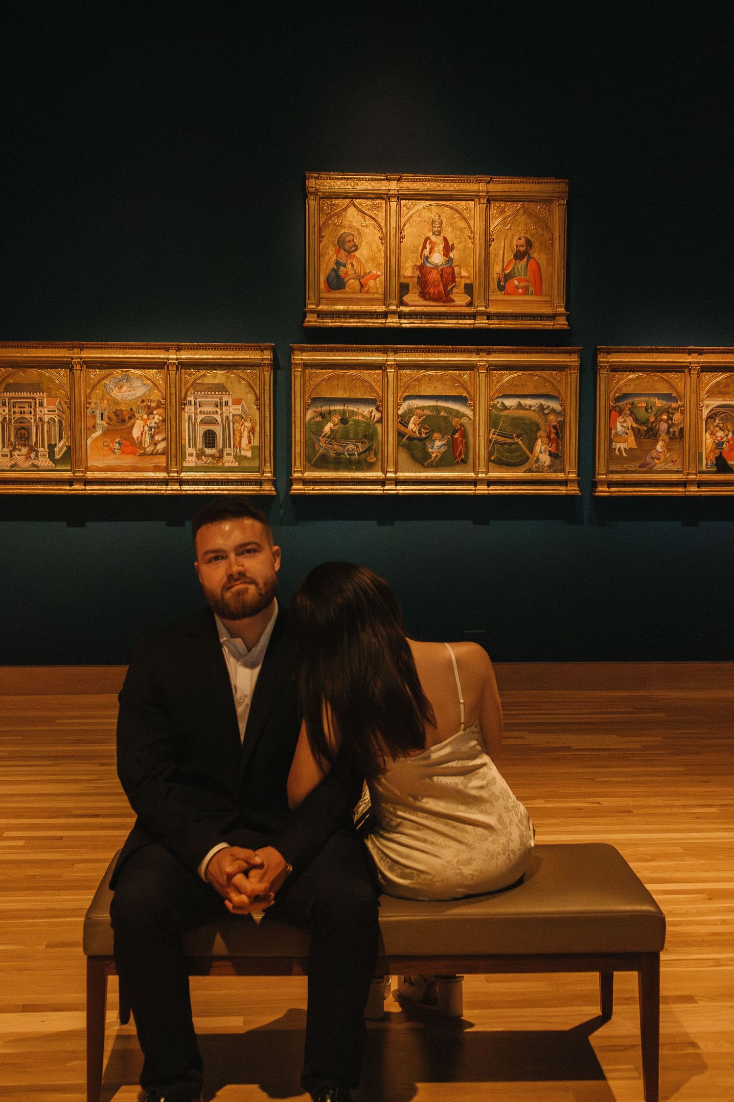 Museum gallery space with golden religious paintings mounted on dark walls and bench seating below.