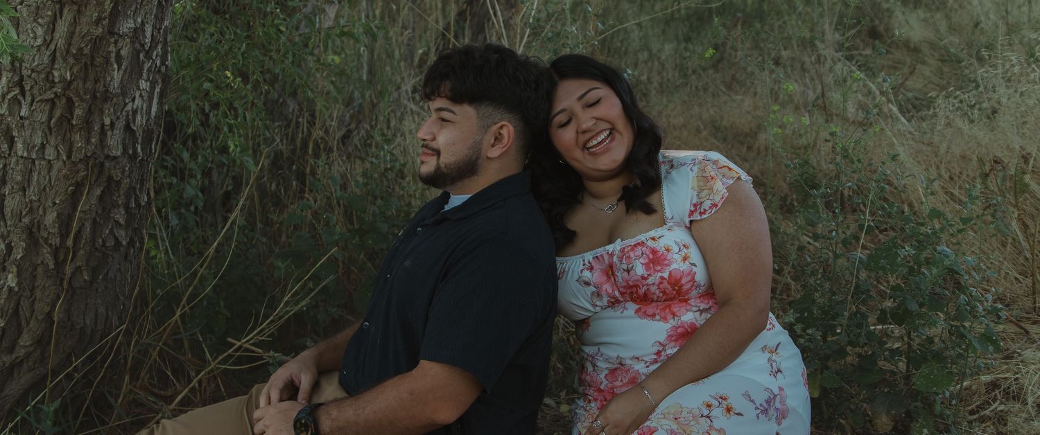 A couple sits together against a tree trunk in a natural outdoor setting.