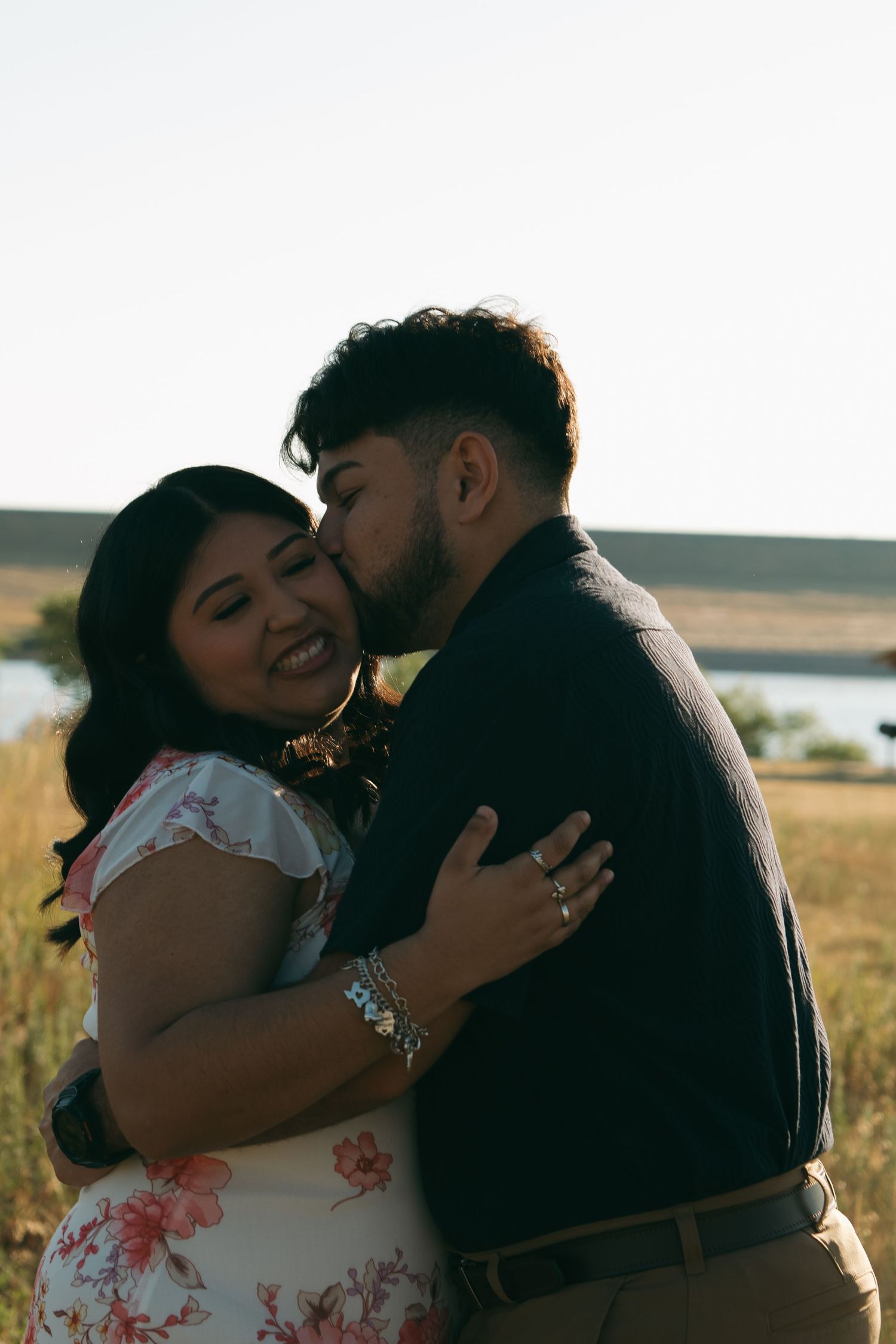 A romantic couple shares an intimate moment in a sunlit field with natural landscape behind them.