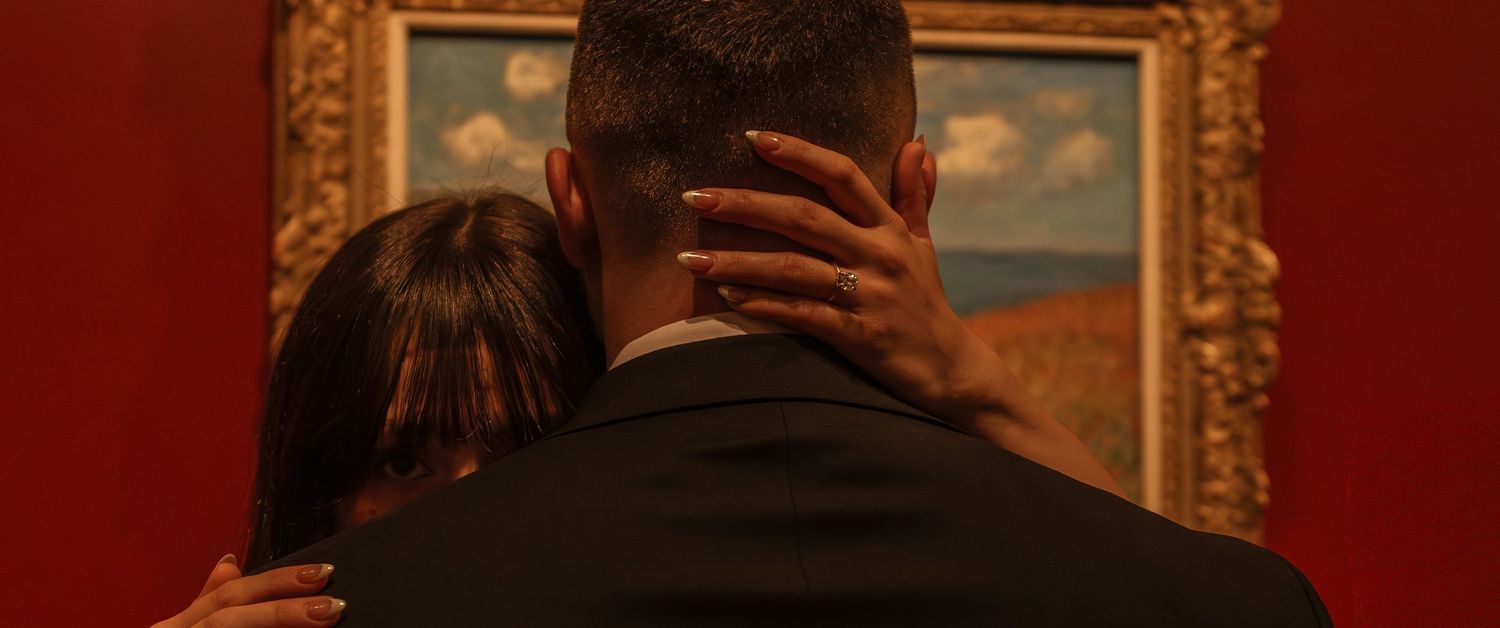 A couple views artwork in an ornate gold frame against a red wall in a dimly lit gallery.